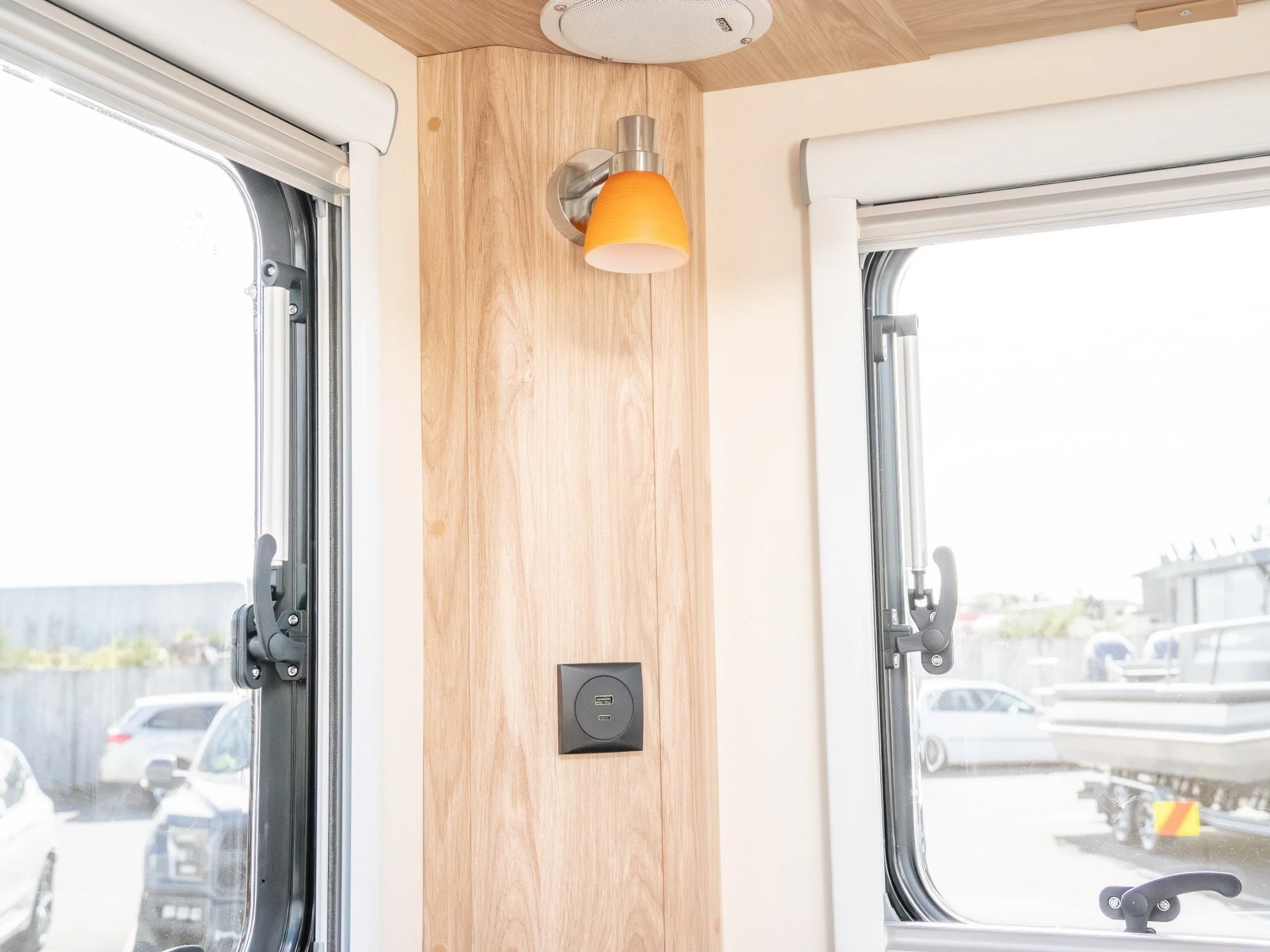 Close-up of a wooden wall panel inside a vehicle, featuring an orange lampshade light, a power outlet, and two window frames on each side with open blinds, showing a parking lot outside.