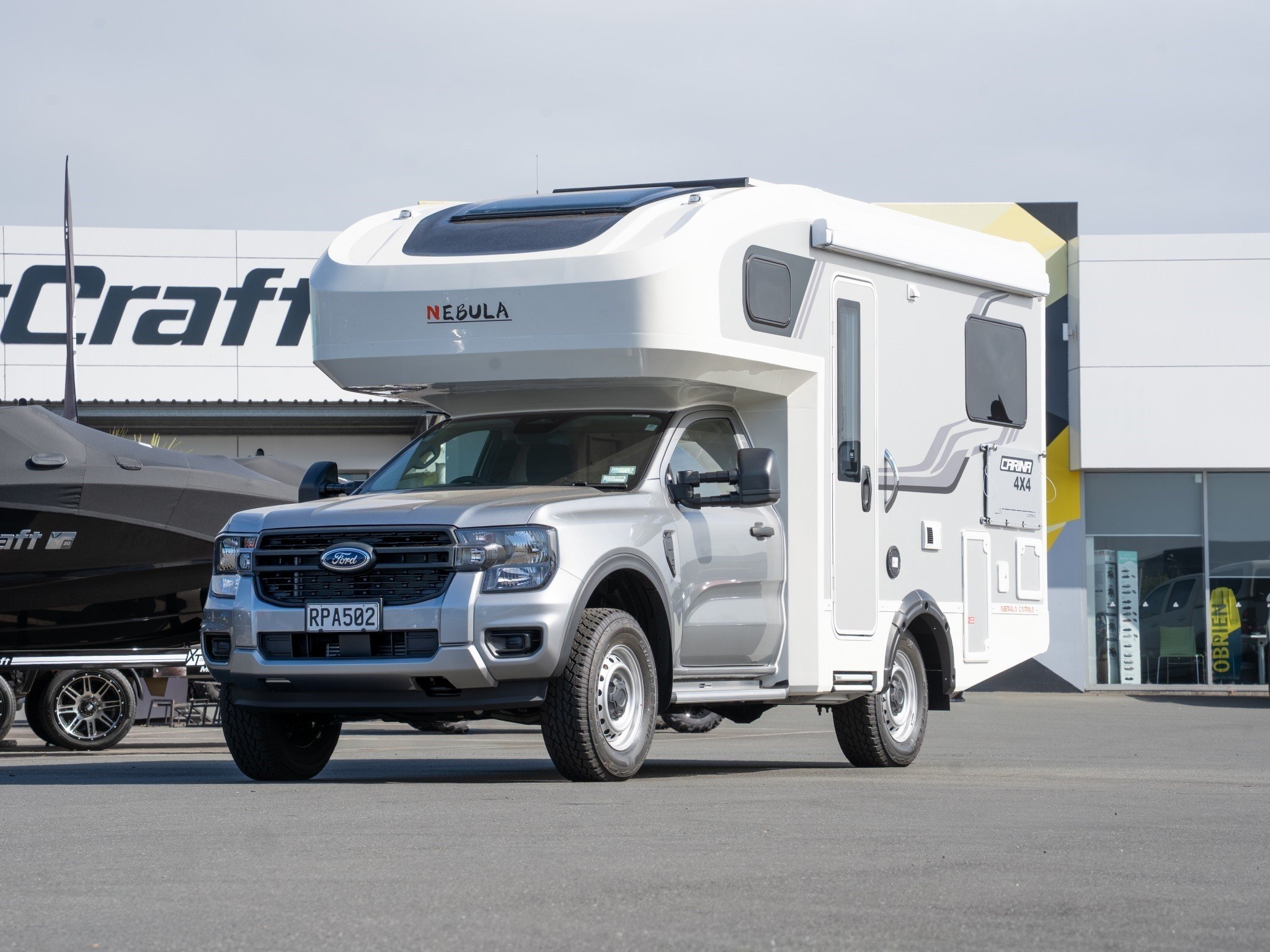 A white motorhome labeled Nebula in a parking lot, with a gray Ford truck attached. The motorhome has windows and compartments, and there are boats and other vehicles nearby. A building with a sign that says Crafts is in the background.