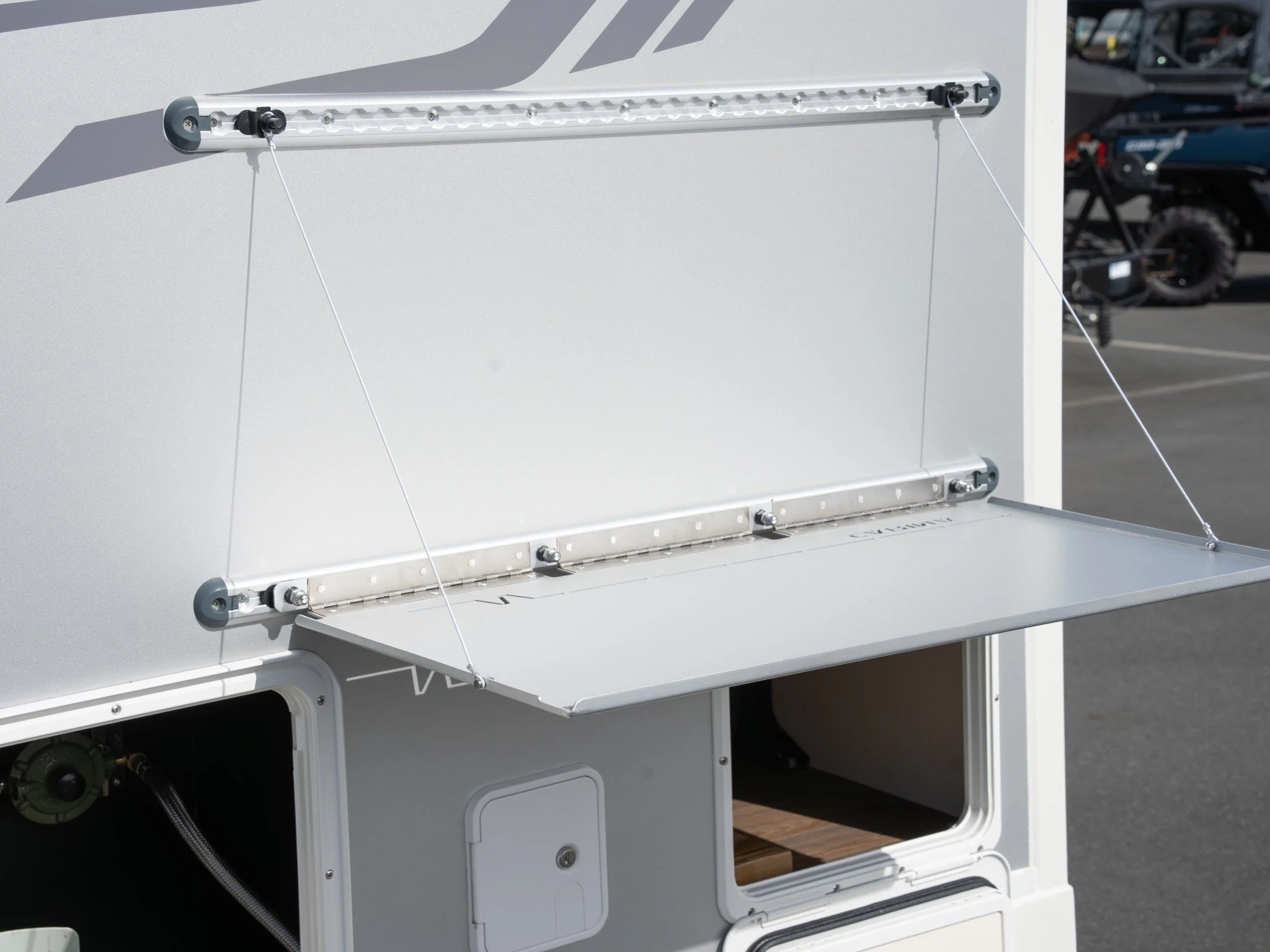 Close-up of a white outdoor cabinet with a fold-down door, metal hinges, and utility wires attached to its side, situated in a parking lot with vehicles in the background.