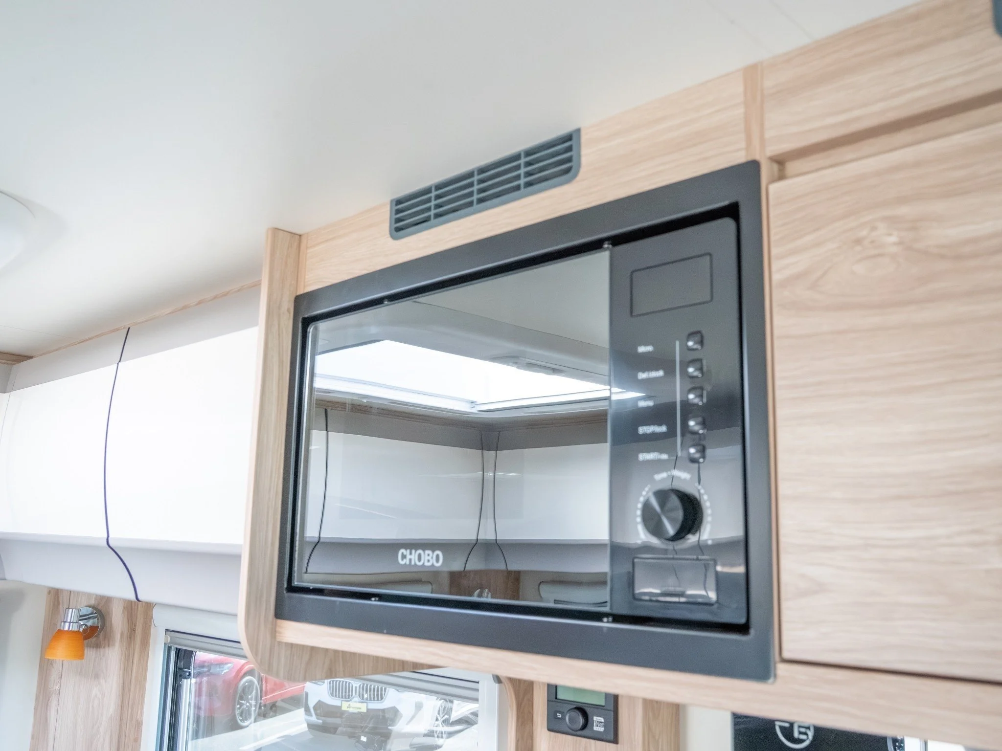 A kitchen with a black microwave oven built into a light wood cabinet, a white countertop, and a window reflecting outside vehicles.