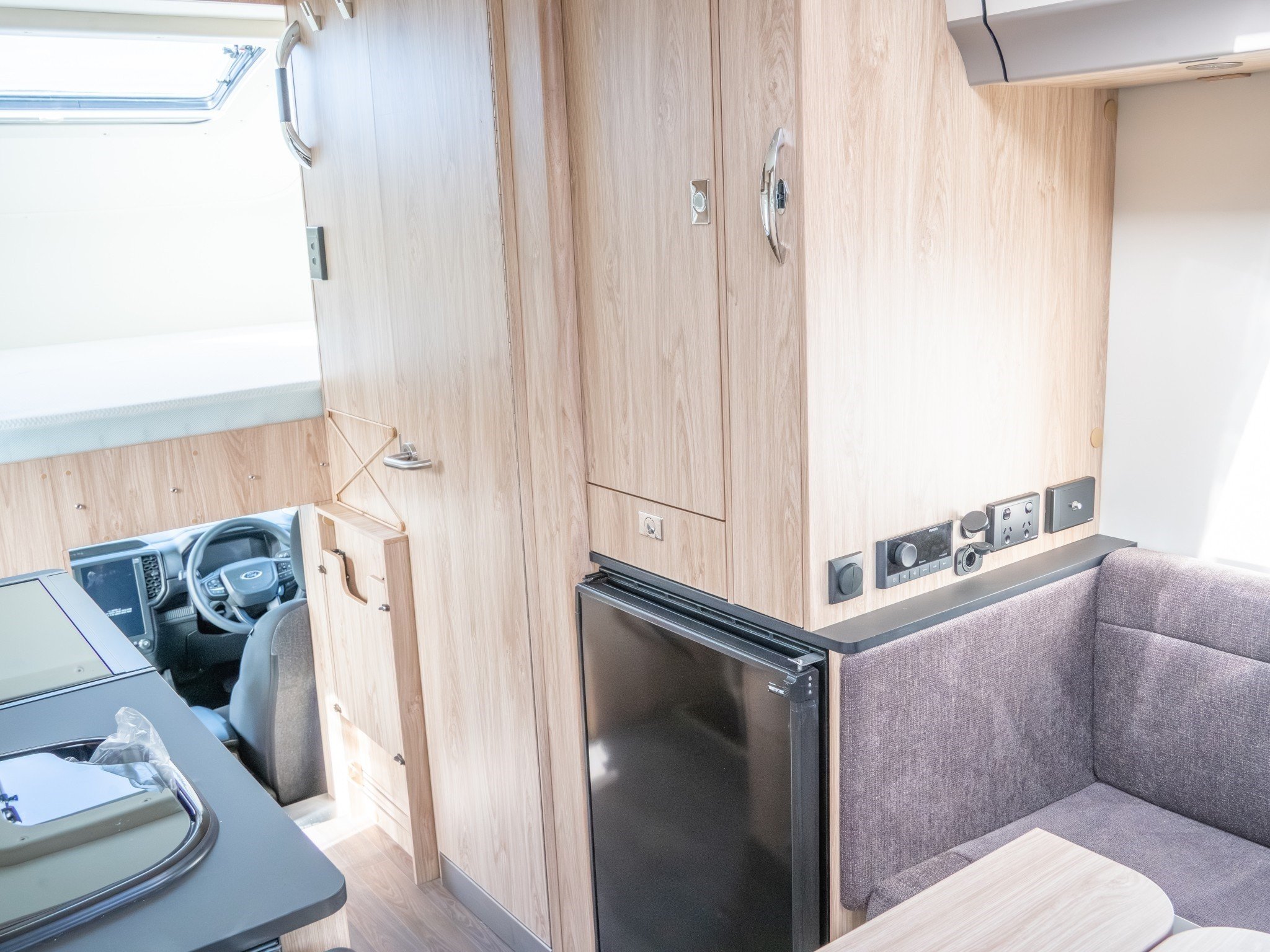 Interior of a tiny home or camper with wooden cabinets, a small black refrigerator, a gray couch, and various electrical outlets and switches on the wall.