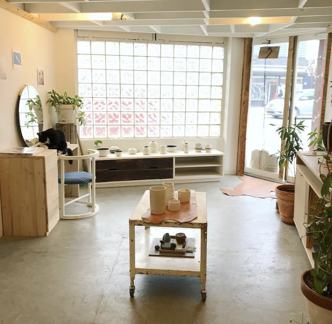 Interior view of a minimalist, well-lit shop or studio with wooden furniture, potted plants, decorative ceramics, and large windows letting in natural light.