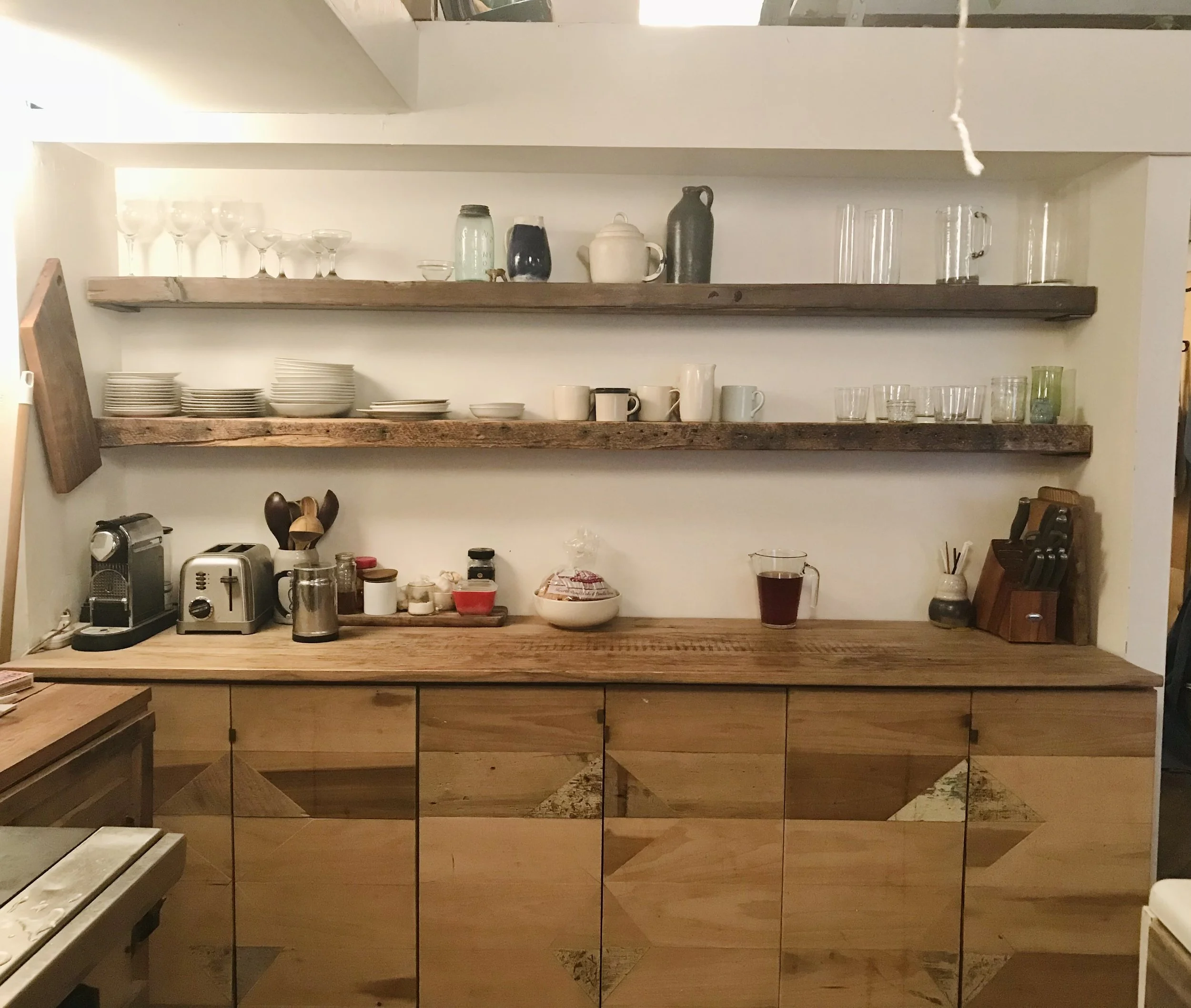 Wooden kitchen cabinet with open shelves holding glassware, plates, cups, and decorative items. Kitchen countertop with coffee machine, toaster, jars, bowl, glass pitcher of dark liquid, and kitchen utensils.