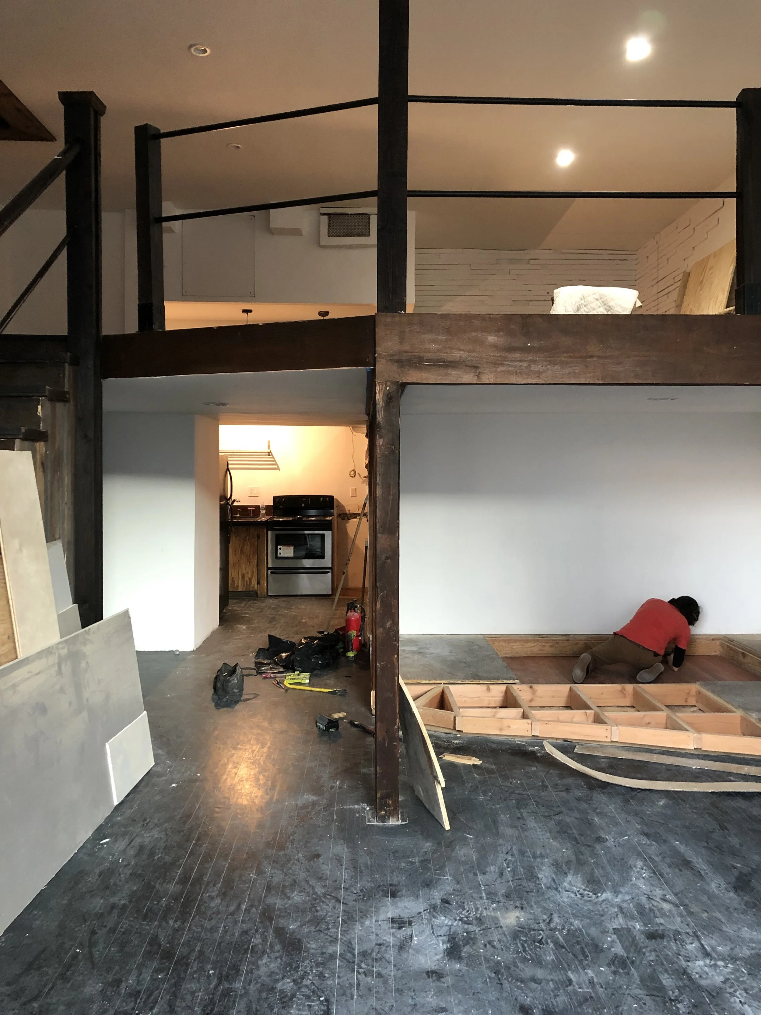 A person working on a home renovation, installing floorboards on a wooden frame, with construction tools and debris on the dark wooden floor.