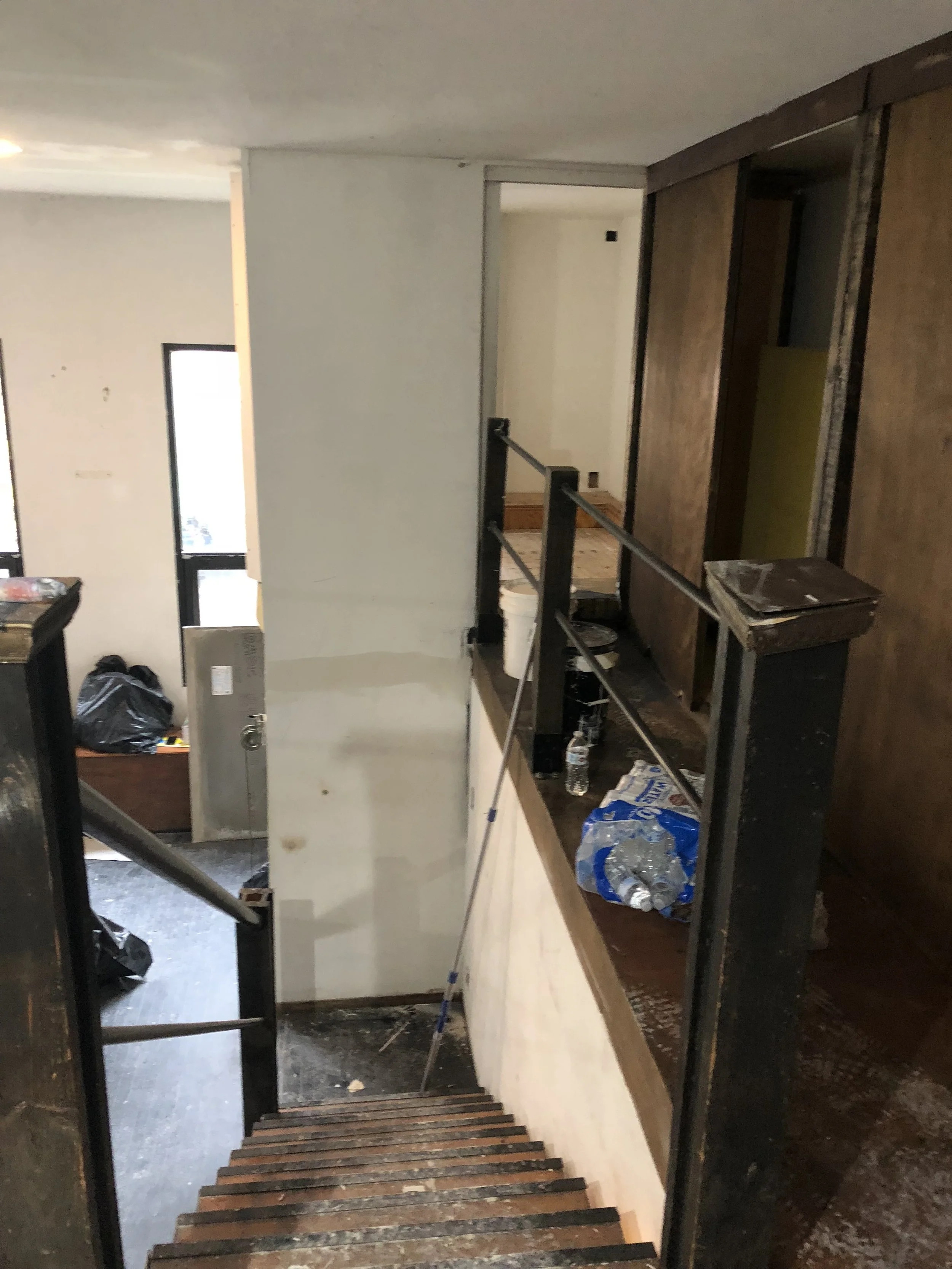 Interior of a house in renovation, showing wooden stairs leading down to a cluttered area with construction materials and tools, partially unfinished walls, and black railings.