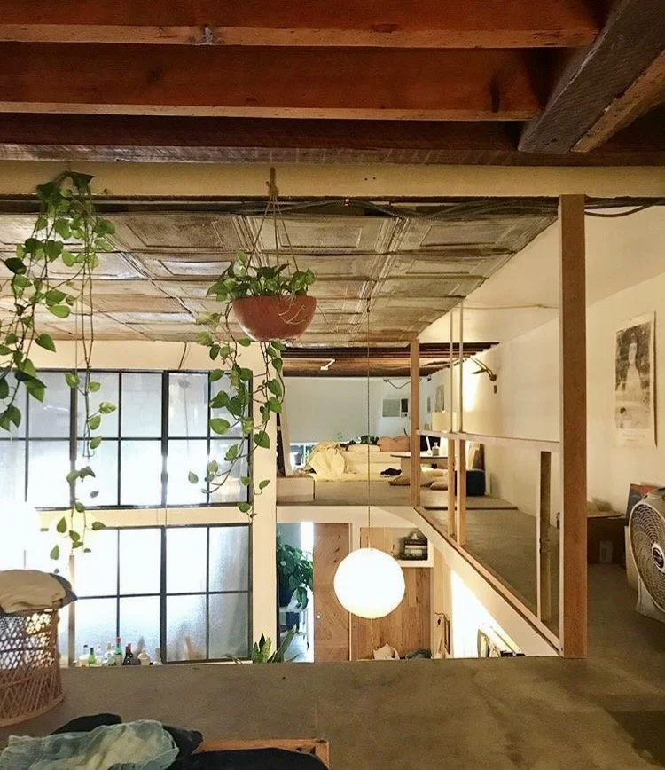 Interior view of a two-story room with hanging plants, a large window, wooden ceiling, and a loft area. The lower level includes a round pendant light and various furniture, while the upper level has a bed and a small sitting area.