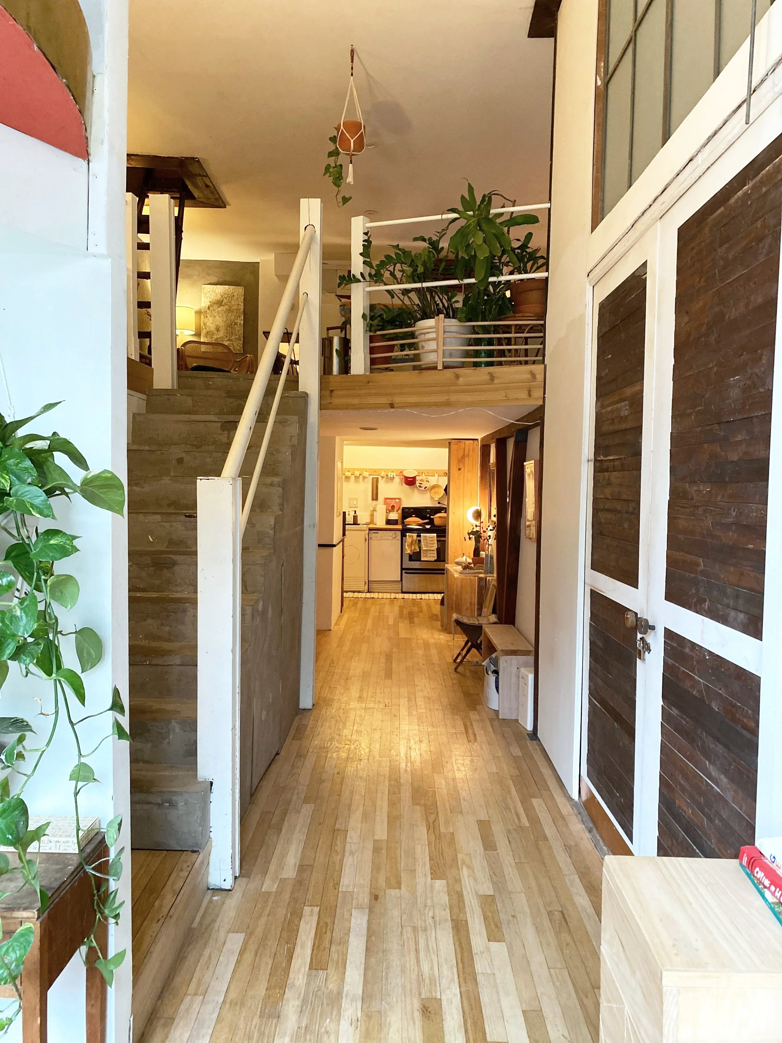 Indoor view of a home with a hallway leading to a kitchen area, filled with wooden and white elements, and various potted plants and furniture.