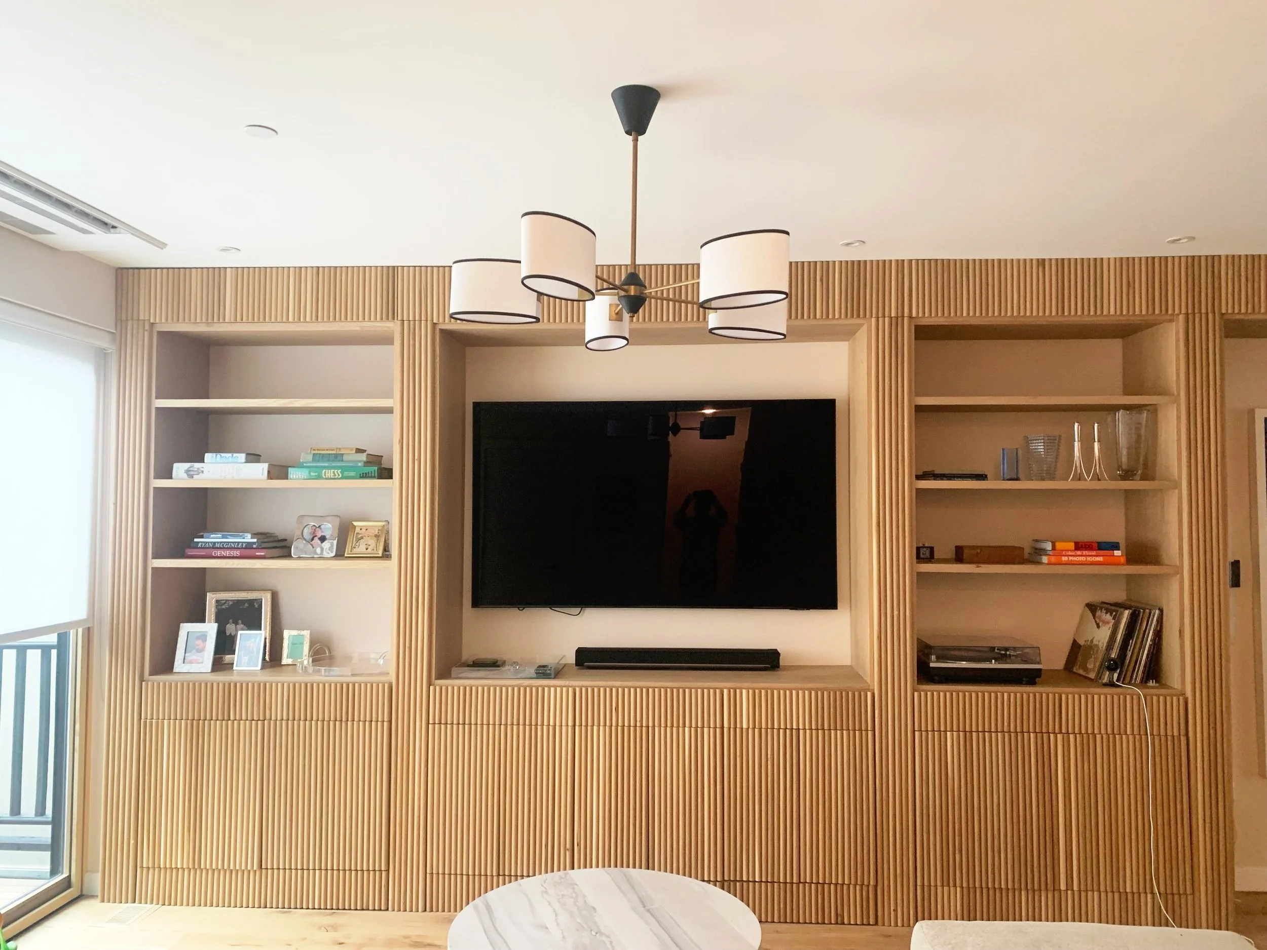 Living room with wooden wall unit, black flat-screen TV, and decorative shelves, with a white ceiling and modern ceiling light fixture.