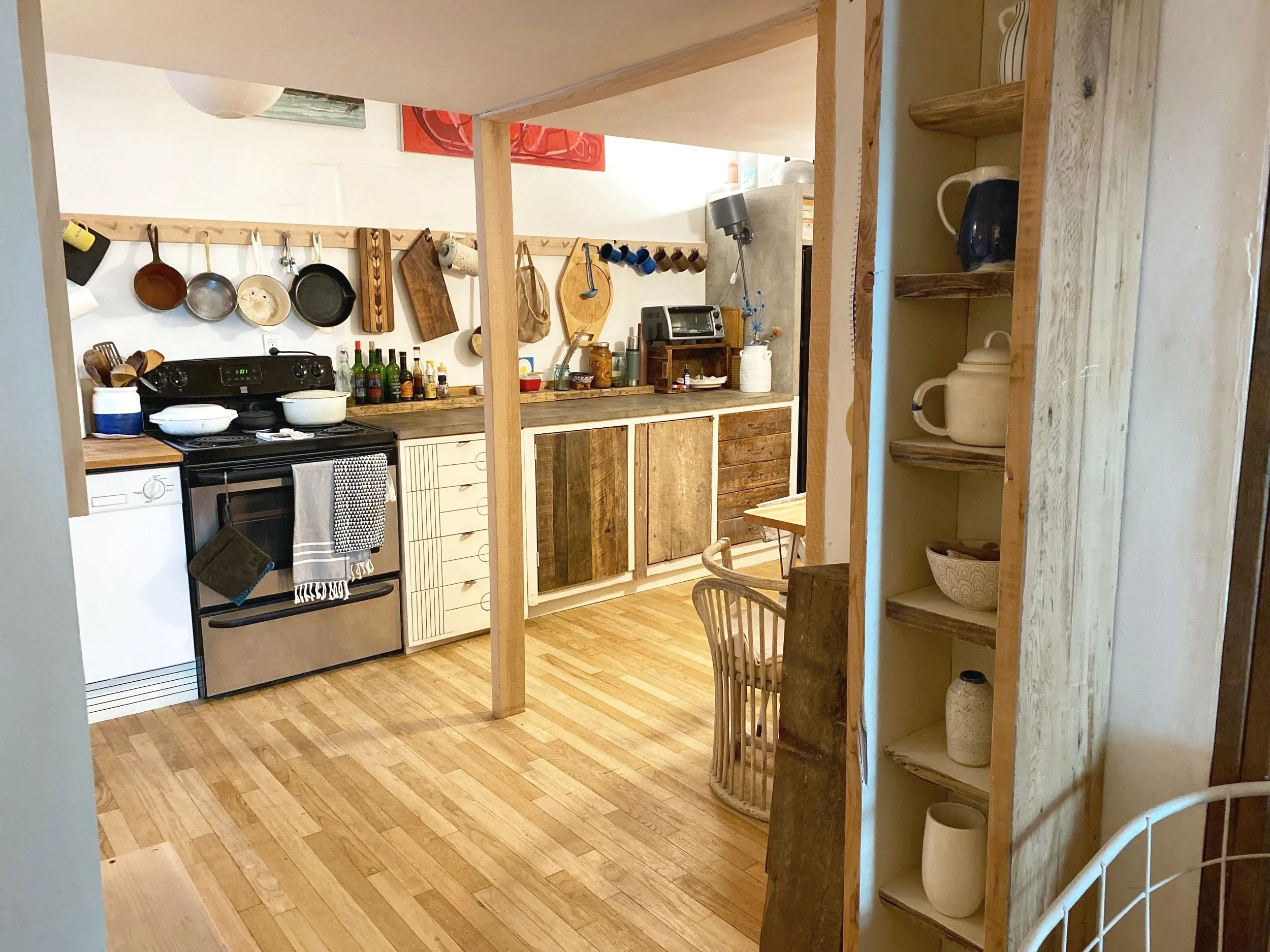 Kitchen with wooden cabinets, hanging pans, a stove, a small table, and open shelving with dishes and bowls.