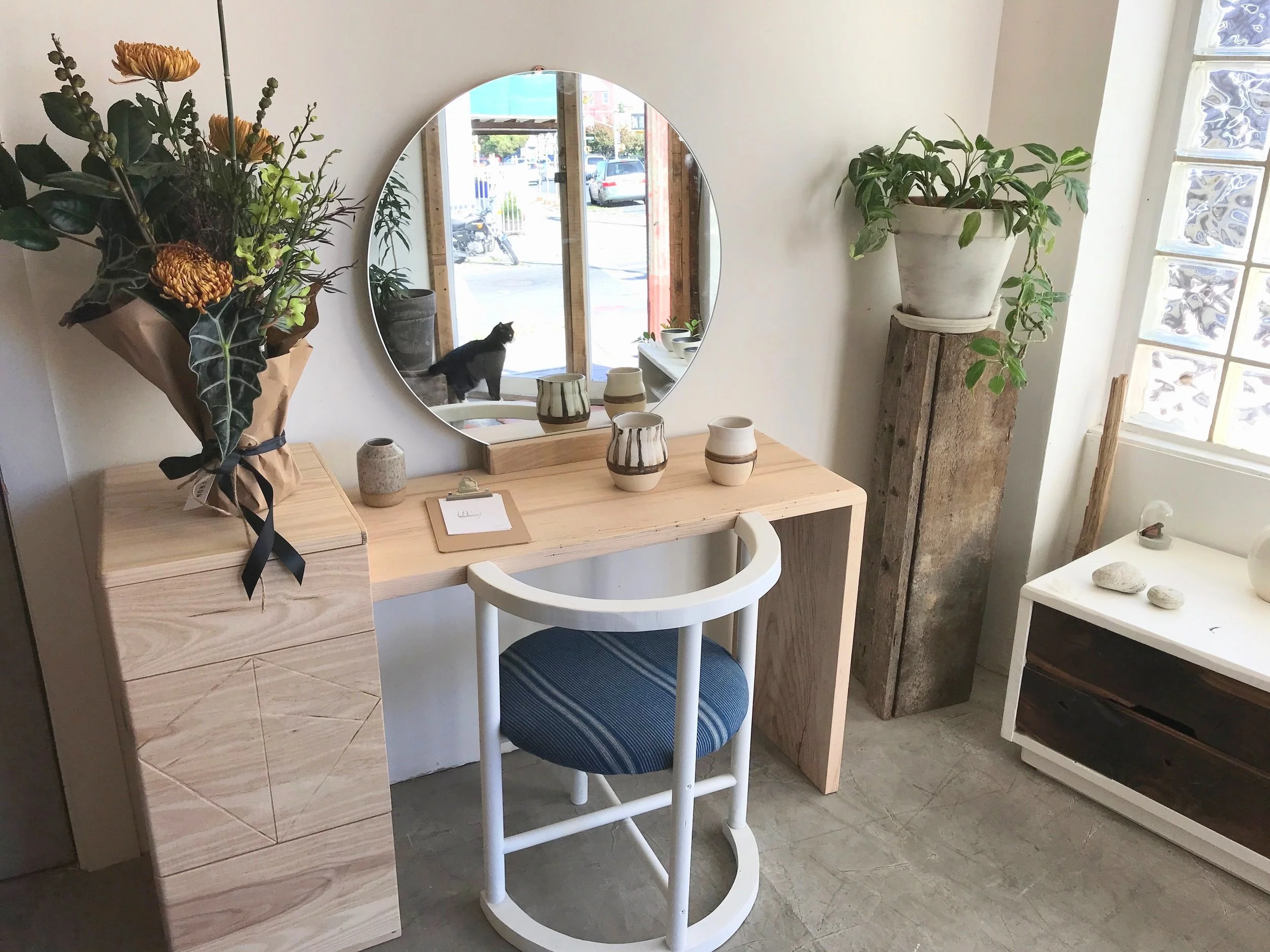 Interior of a minimalistic room showing a wooden vanity with a round mirror, a white chair with a blue cushion, a bouquet of flowers on a wooden cabinet, potted plants, and some decorative stones on a white table. Through the mirror, a cat is seen sitting by the window outside.