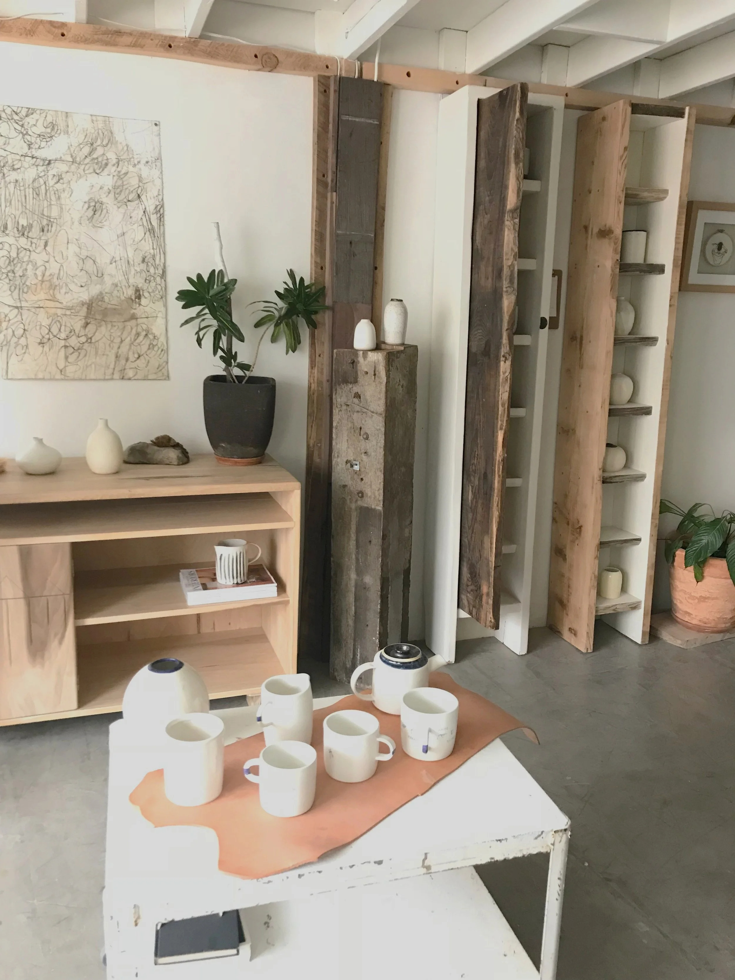 Interior of a home decor store featuring ceramic mugs and vases on a white distressed table, wooden shelves with vases and decorative items, and potted plants against a white wall with abstract artwork.
