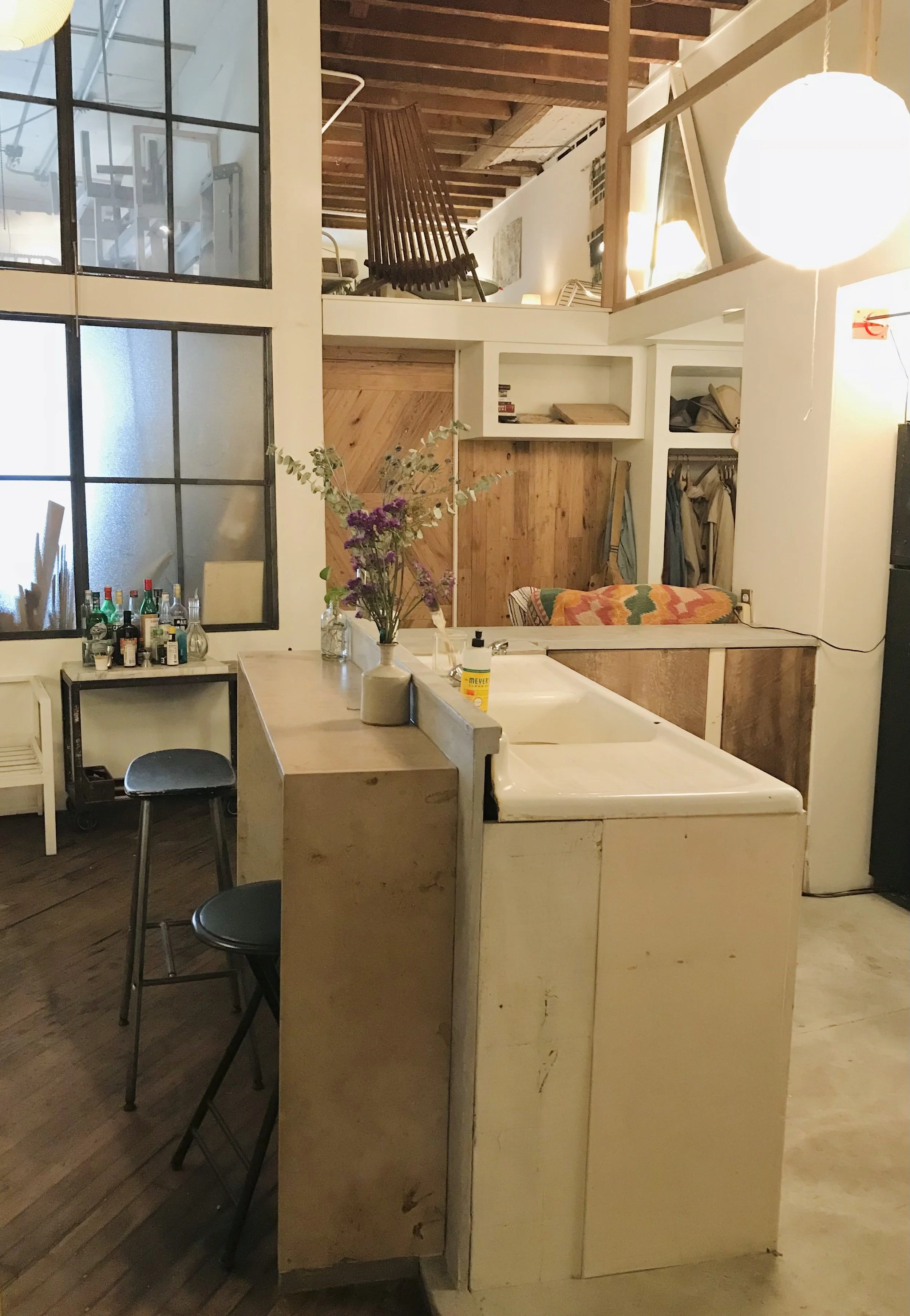 Interior view of a cozy, rustic apartment with an open kitchen and lofted bedroom. Features include a kitchen island with a vase of flowers, bar stools, a bar cart with bottles, a large frosted window, and warm wood accents.