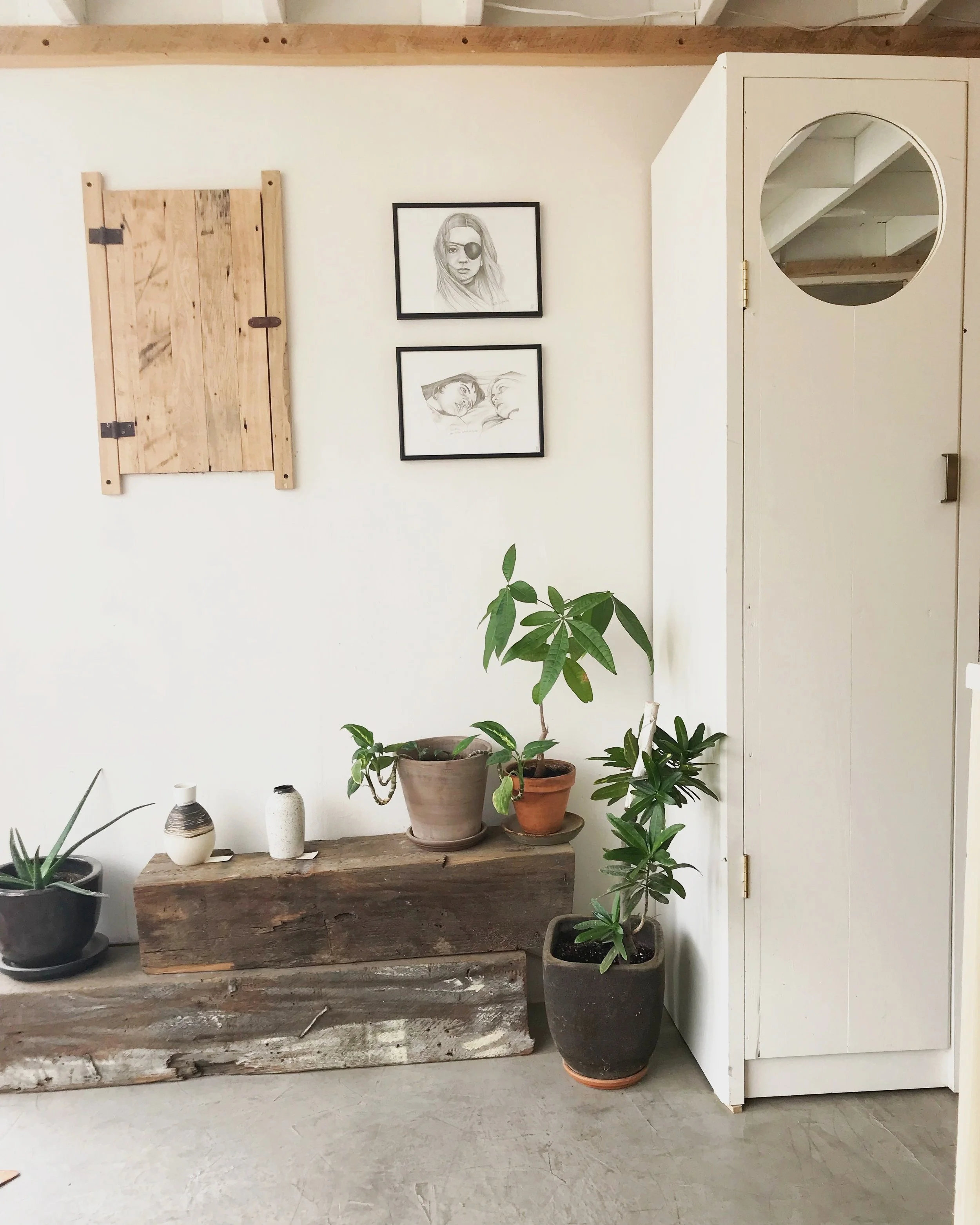 Interior view of a room with a white wall decorated with two framed black and white drawings of women, a wooden vent cover, and a white cabinet with a round mirror. Several potted plants sit on a wooden beam and on the floor.