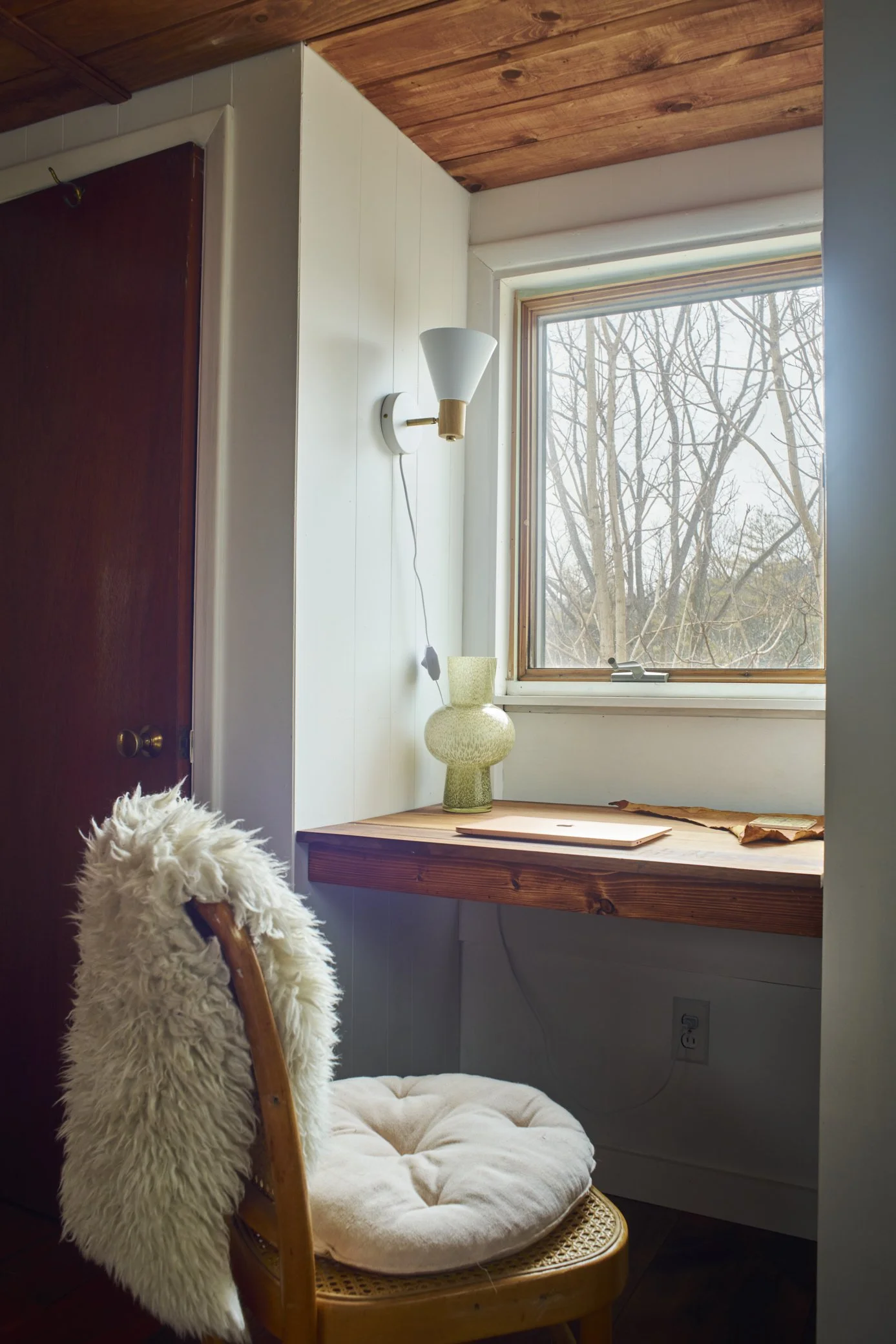 Corner of a room with a wooden desk, a lamp, and a decorative vase by a window showing leafless trees. There is a cozy chair with a sheepskin throw and a cushion.