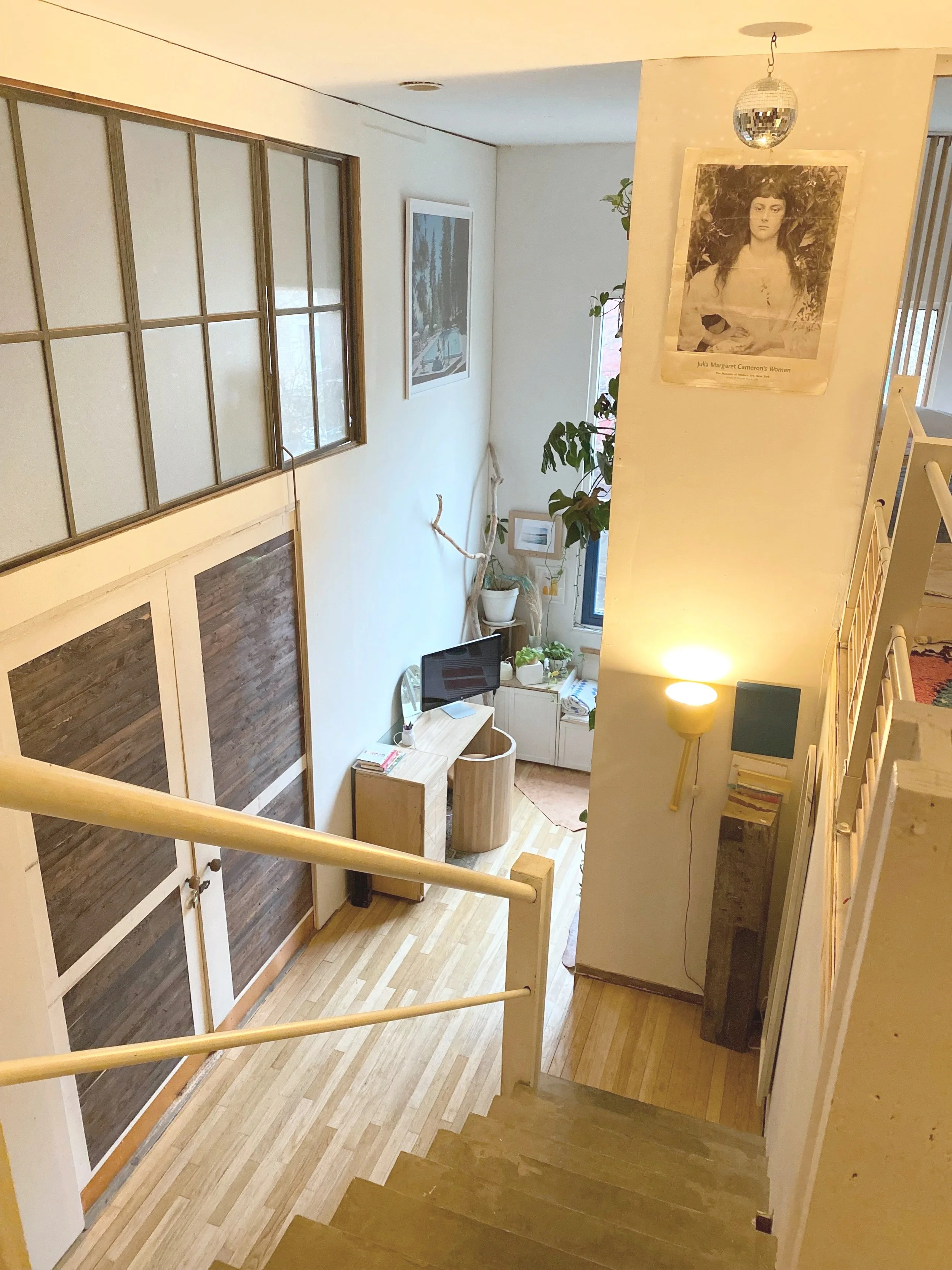 View from the staircase down to the living area with a small desk, television, green plants, and framed artwork.