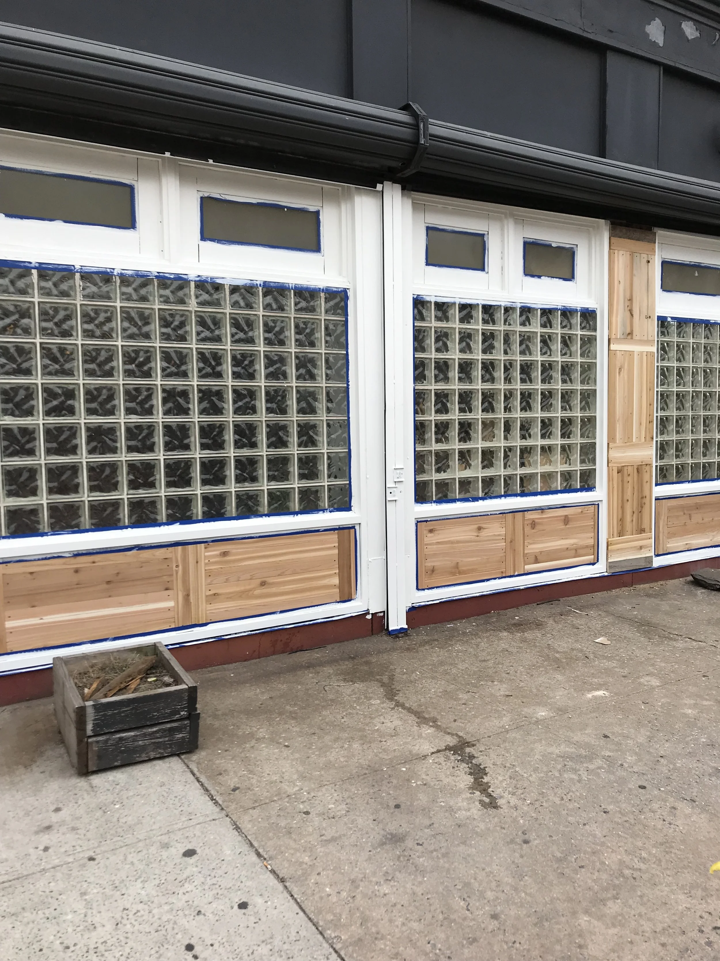 A storefront with large glass block windows and white framing, partially covered with new wood paneling, on a concrete sidewalk.