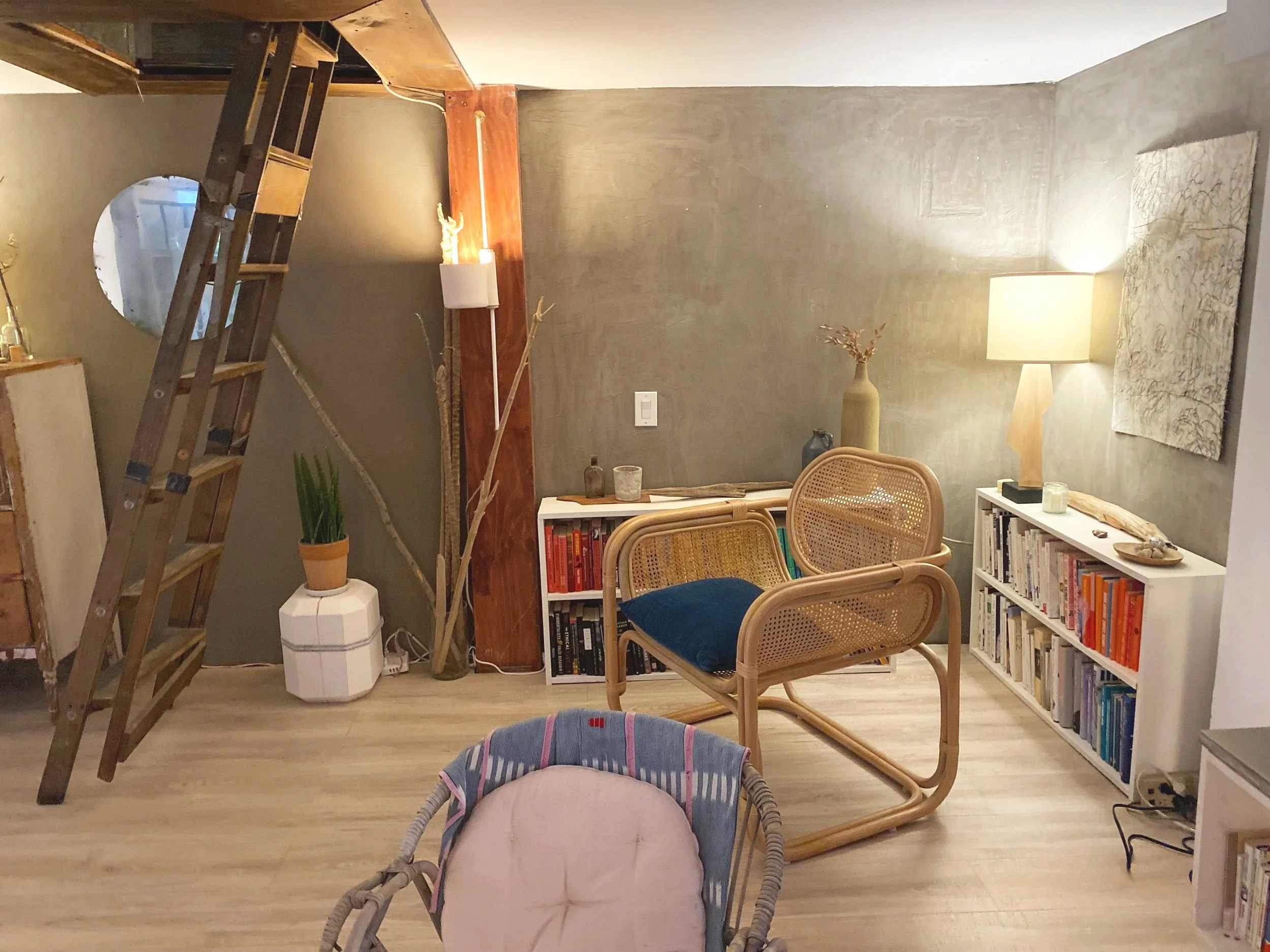 Living room with wooden ladder, bookshelf, chairs, plants, and a lamp, visible on a tan wood floor and textured wall.