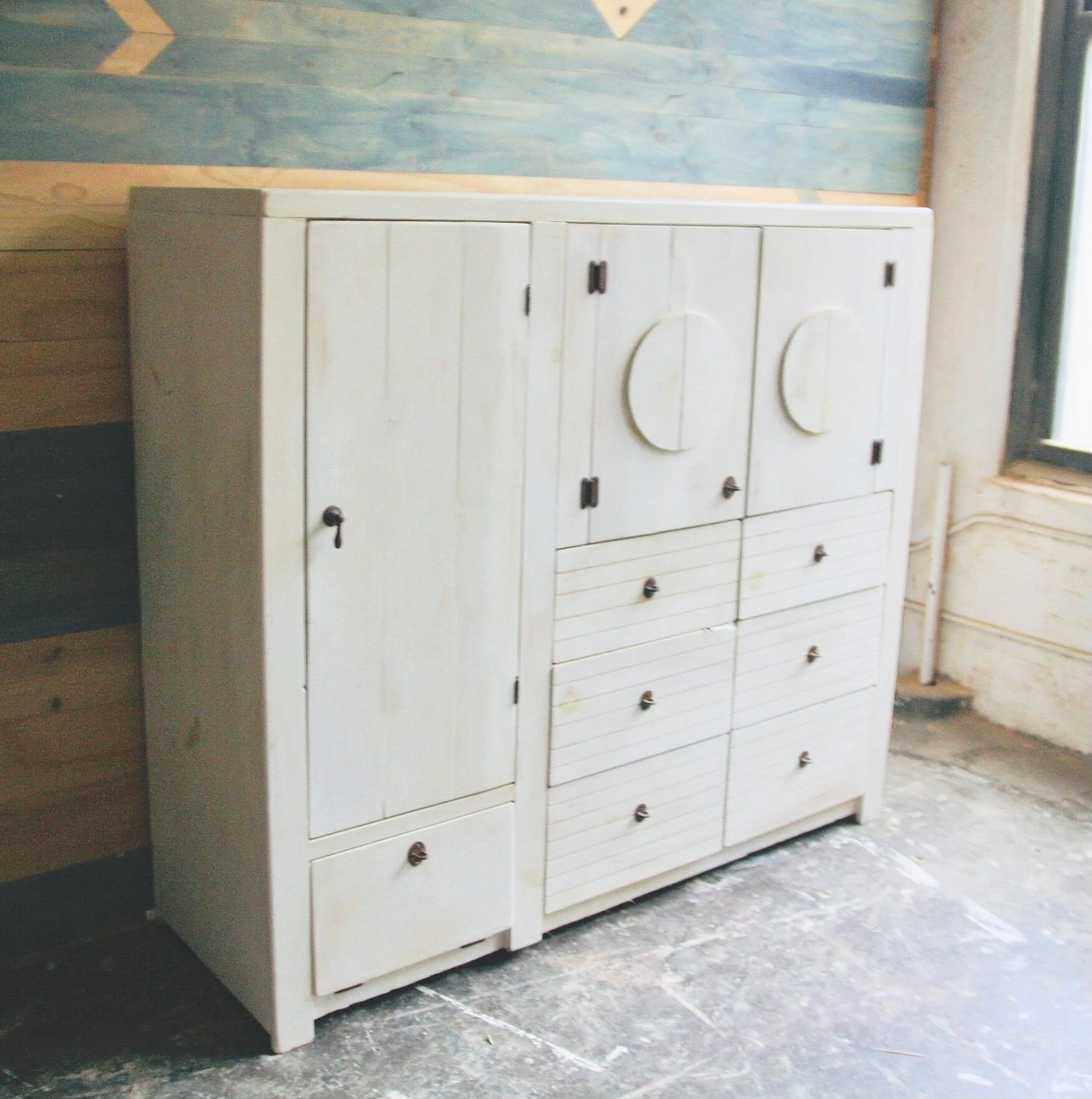 White wooden cabinet with multiple doors and drawers, some with black handles, in a room with a wooden wall and a window nearby.