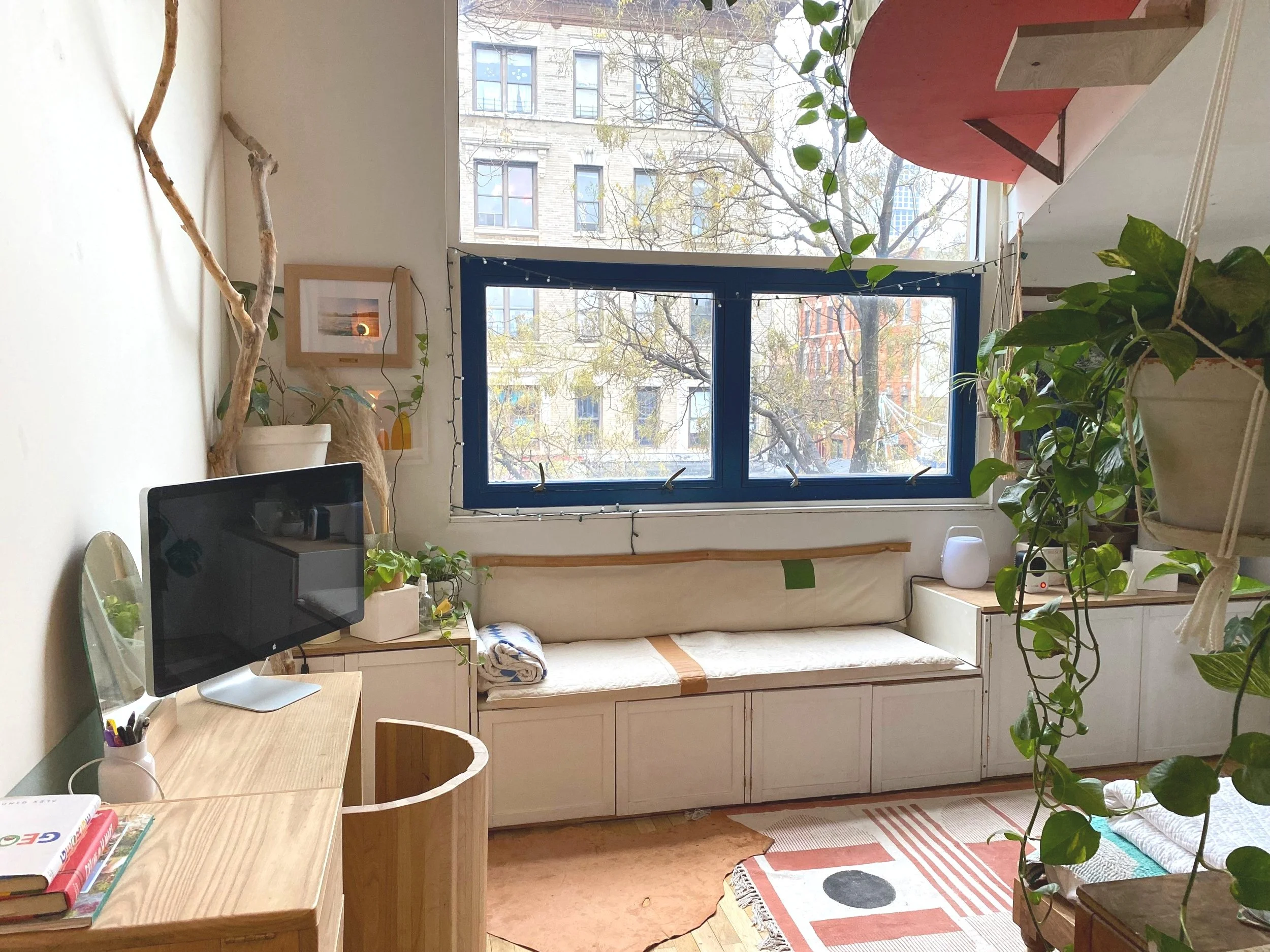 Cozy living room with white built-in window seat, a large window with city view, hanging plant, small TV on a desk, and various potted plants.