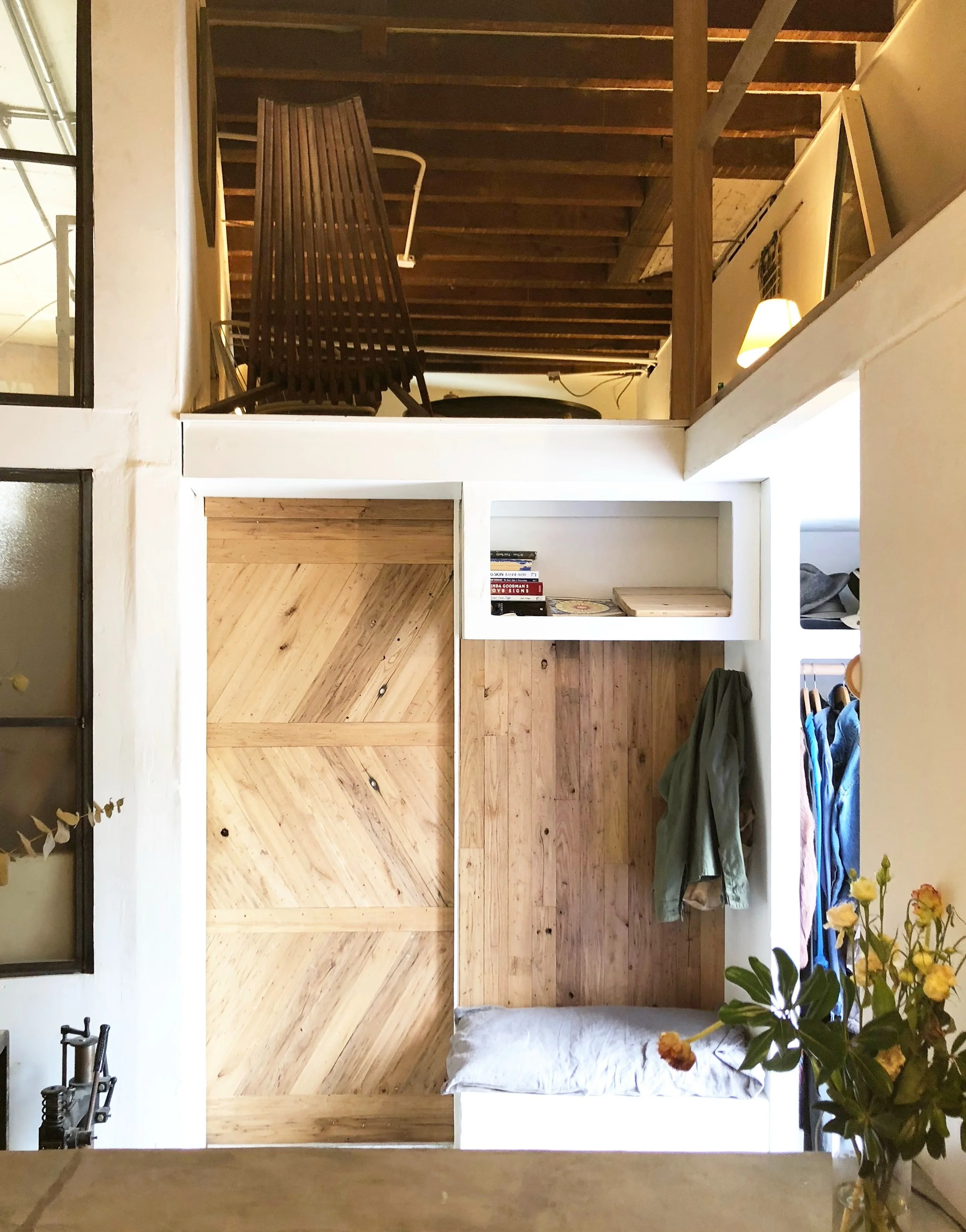 Interior of a cozy living space with a wooden nook featuring a pillow, hanging clothing, and a small shelf with books, adjacent to a modern industrial-style window and a nearby vase with yellow flowers.
