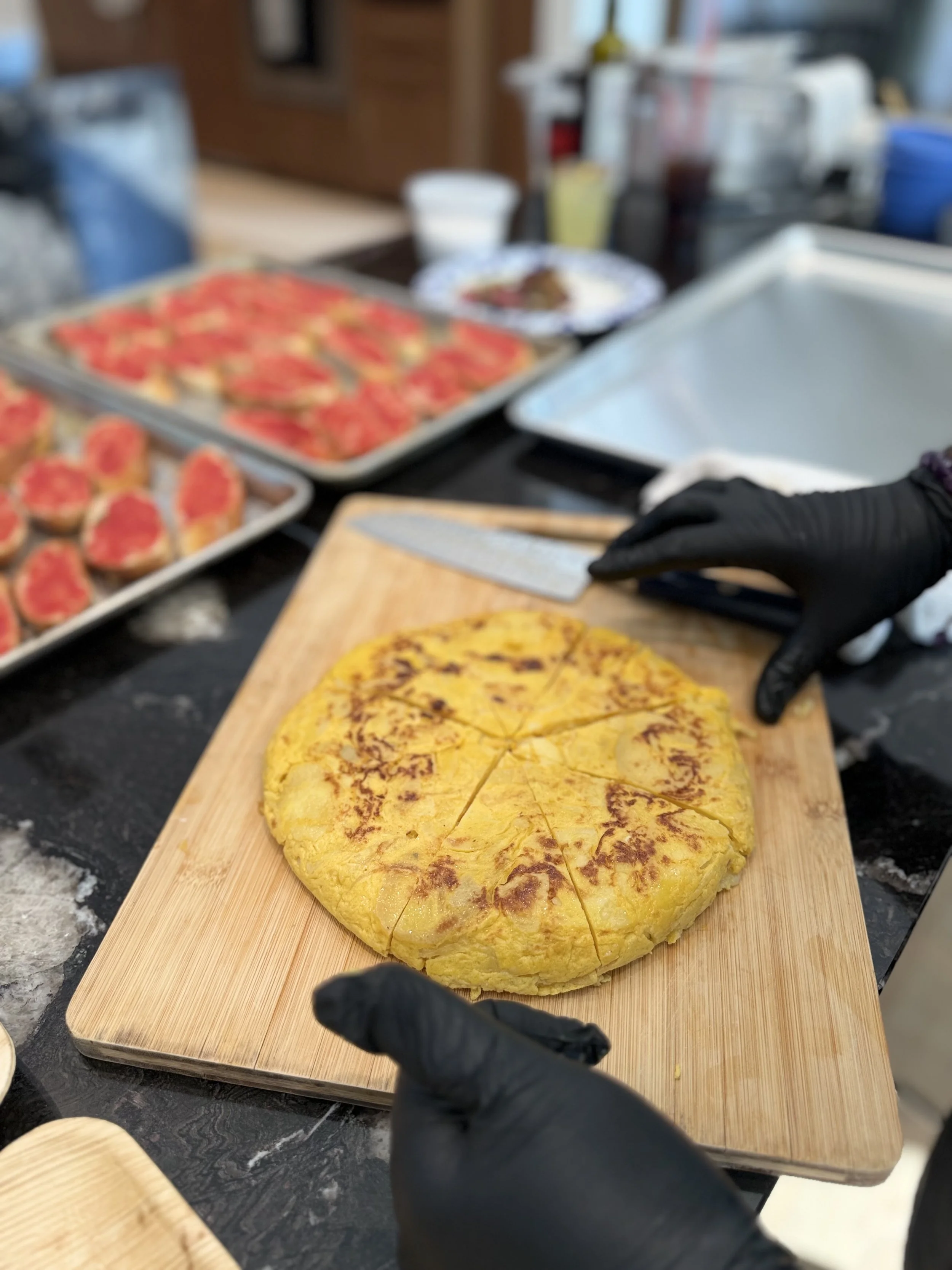 A person wearing black gloves is slicing a round, yellow-colored dish on a wooden cutting board. In the background, there are trays of food, including sliced tomatoes and other dishes, with a blurred kitchen setting.