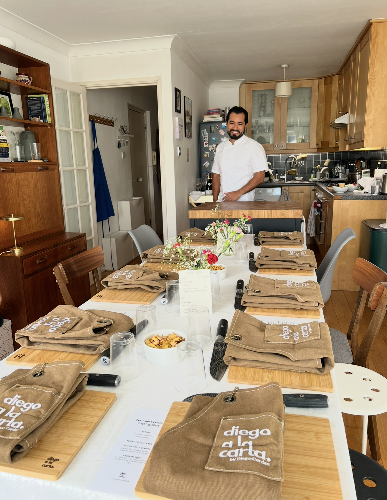 A man in a white chef's coat standing in a kitchen behind a dining table set with brown cloth napkins, cutting boards, and glasses, with a dining room decorated with flowers, books, and kitchen appliances.