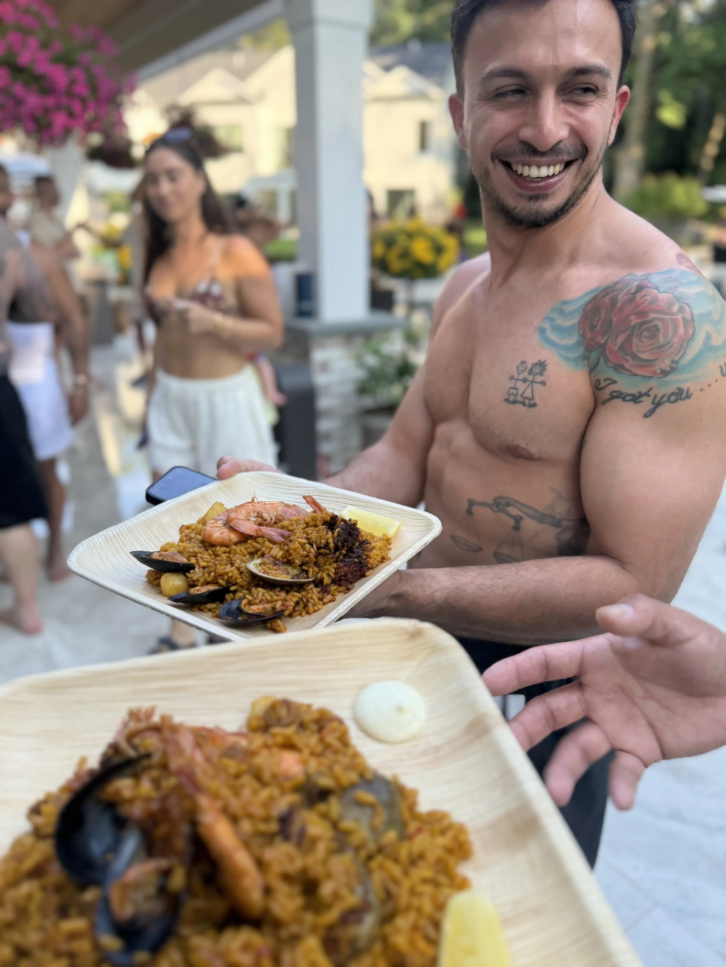 A shirtless man with tattoos on his arm and chest smiling while holding a plate of paella, with other people in the background at an outdoor gathering.