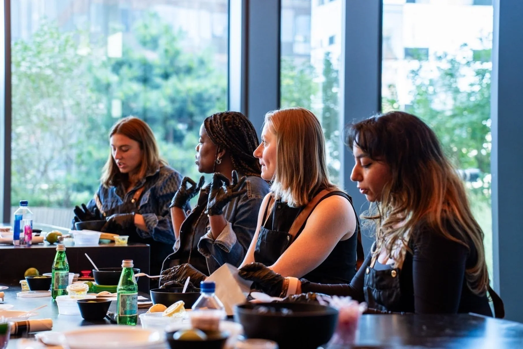 Four women sitting at a table engaged in a cooking or crafting activity with ingredients and supplies on the table in front of them, large windows with greenery outside in the background.