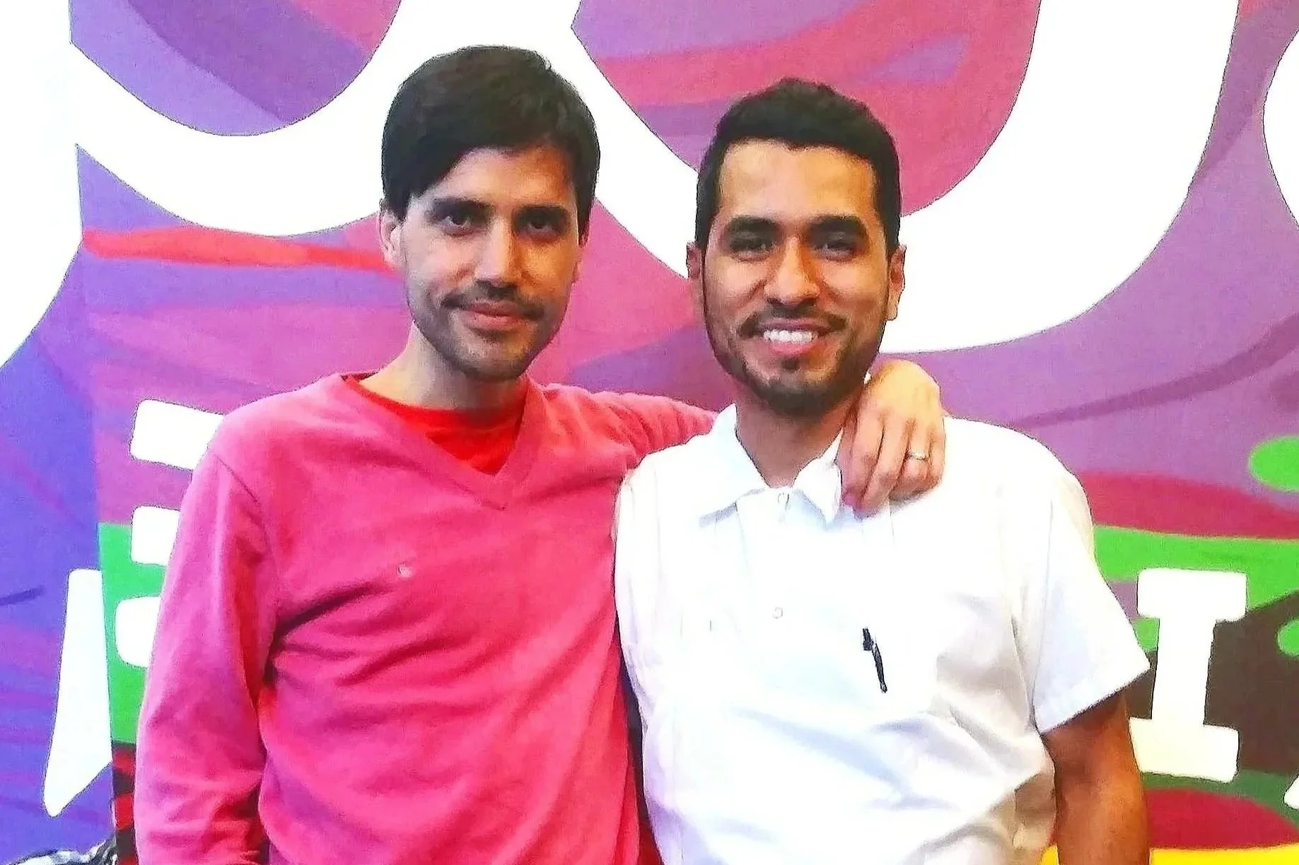 Two men standing close together with their arms around each other, smiling at the camera, with a colorful background.