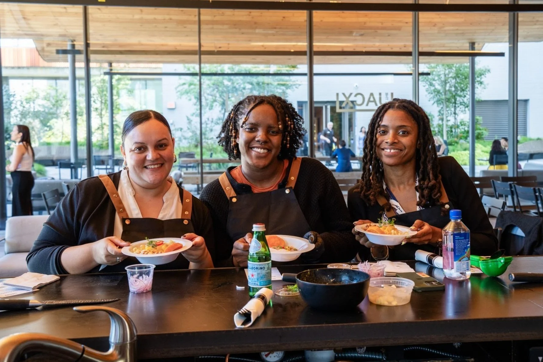 Three women smiling and holding bowls of food at a restaurant table, with a large window and outdoor patio in the background.