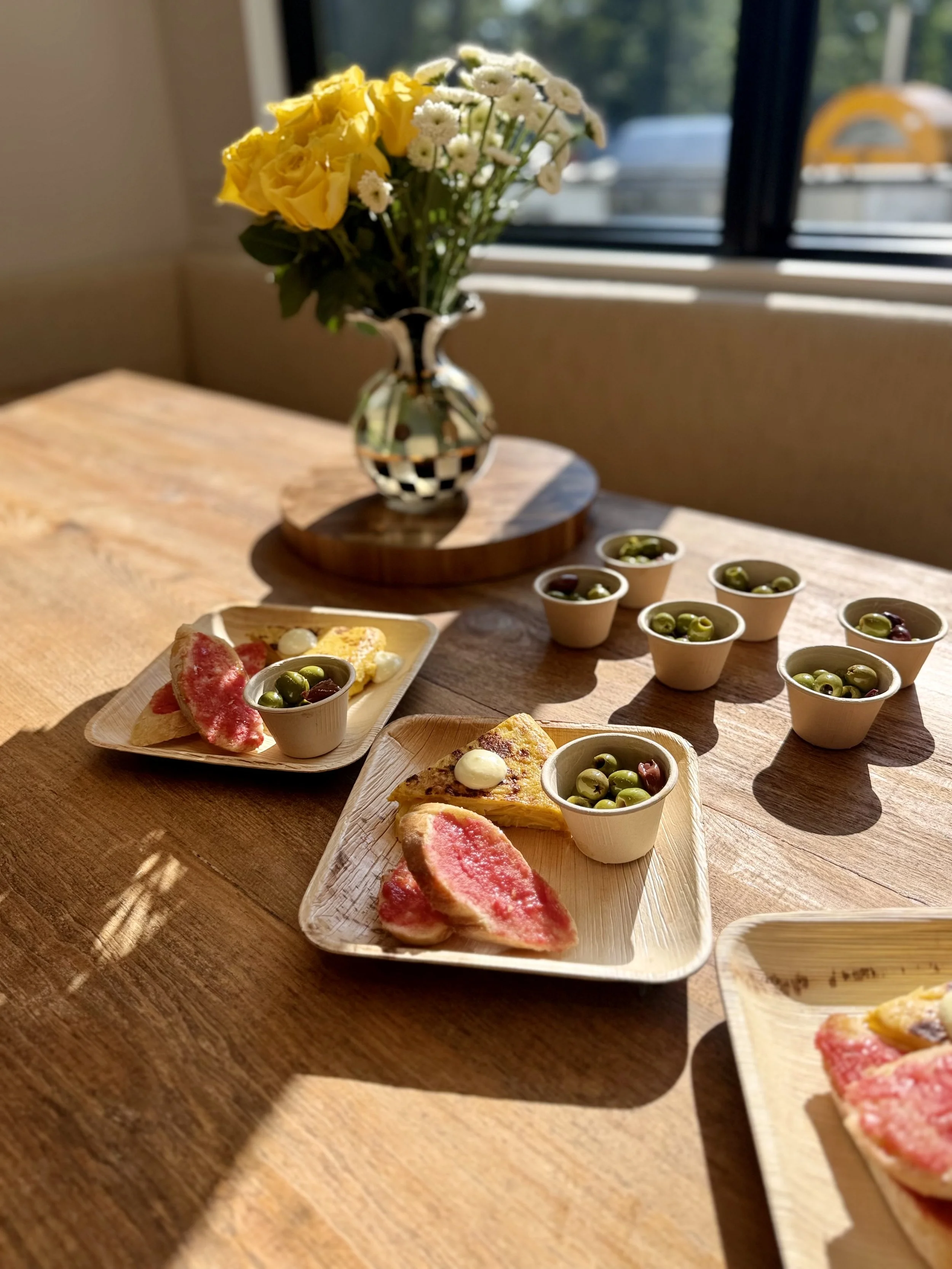 A wooden table with small plates of food, including slices of watermelon, cheese, green olives, and pizza, in front of a window with a vase of yellow and white flowers.