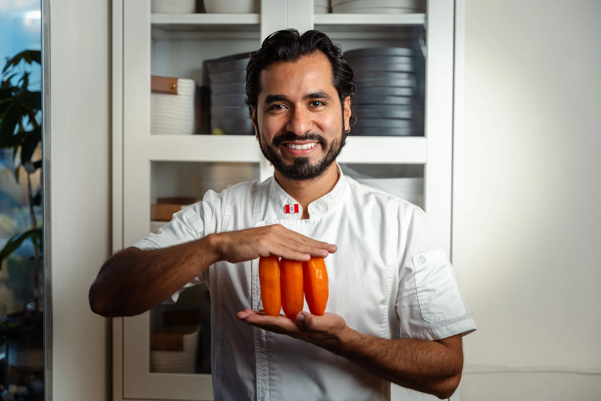 Man with dark hair and beard wearing a white chef jacket holding three carrots, standing in a kitchen with glass-front cabinets and greenery outside.