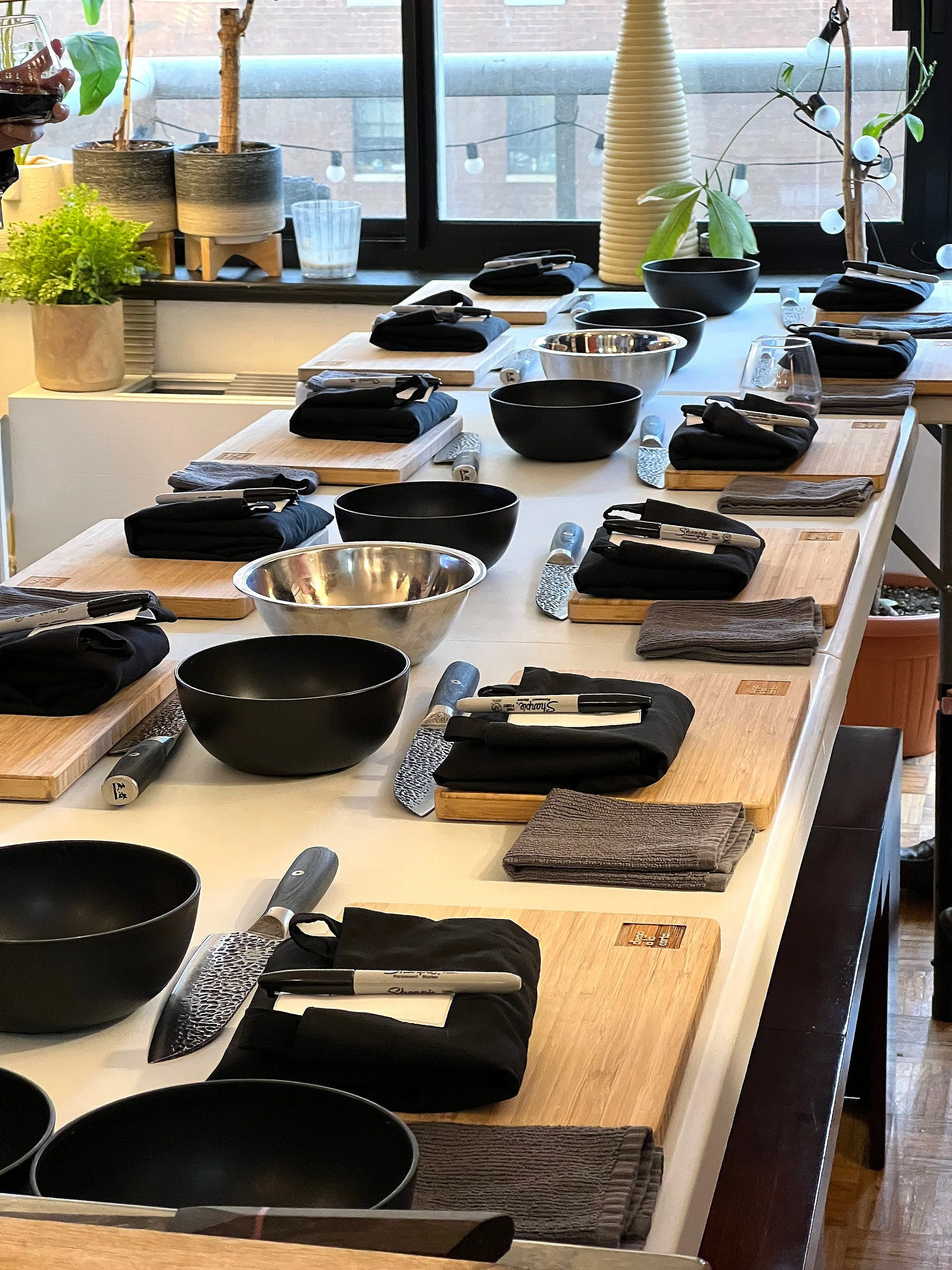 A kitchen counter set up for a cooking class, with black bowls, stainless steel mixing bowls, cutting boards, knives, and black napkins, arranged near a window with plants and string lights in the background.