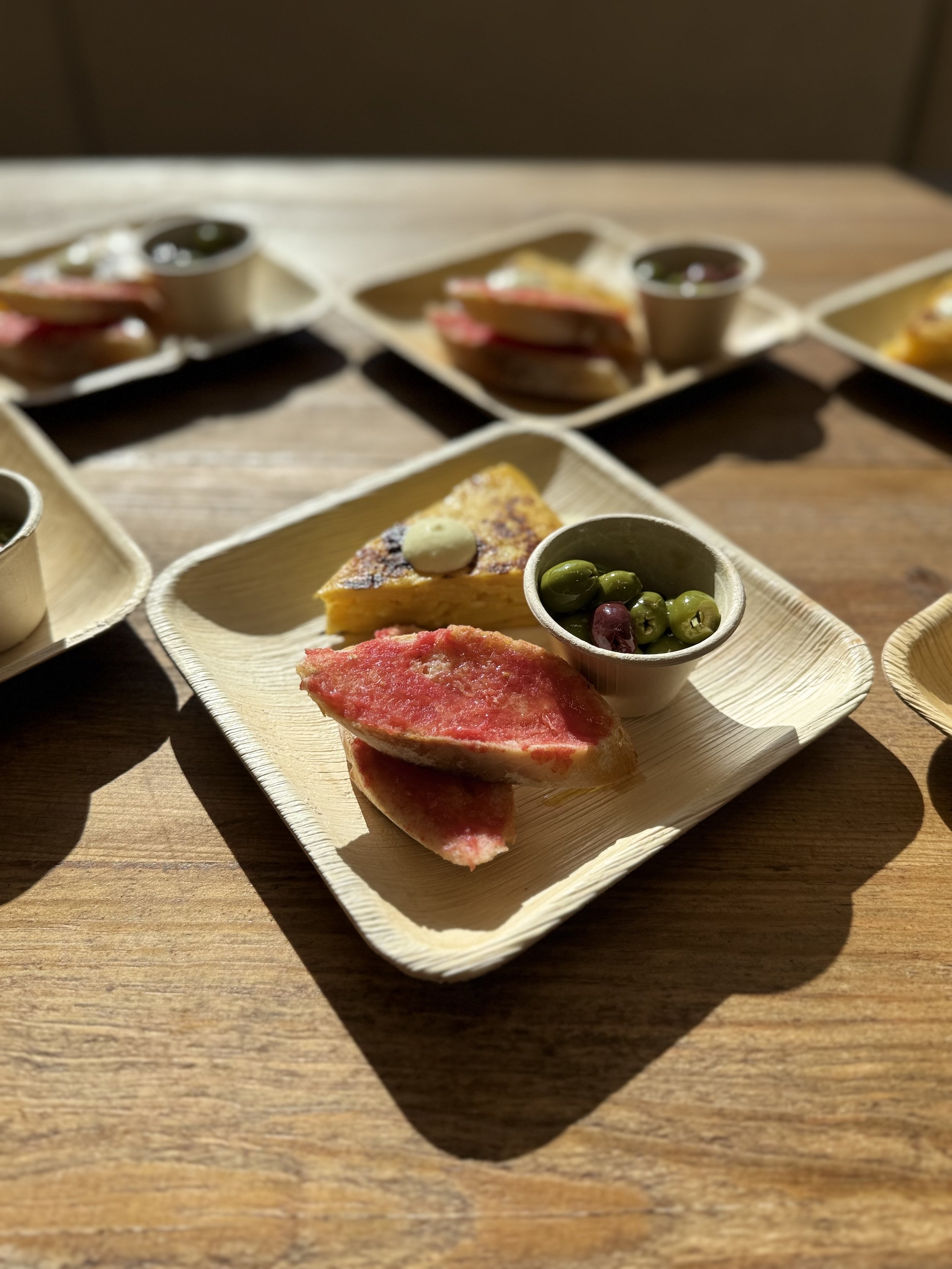 Miniature food items on disposable trays, including slices of cake with dollops of cream, and small cups of green olives, served on a wooden table.