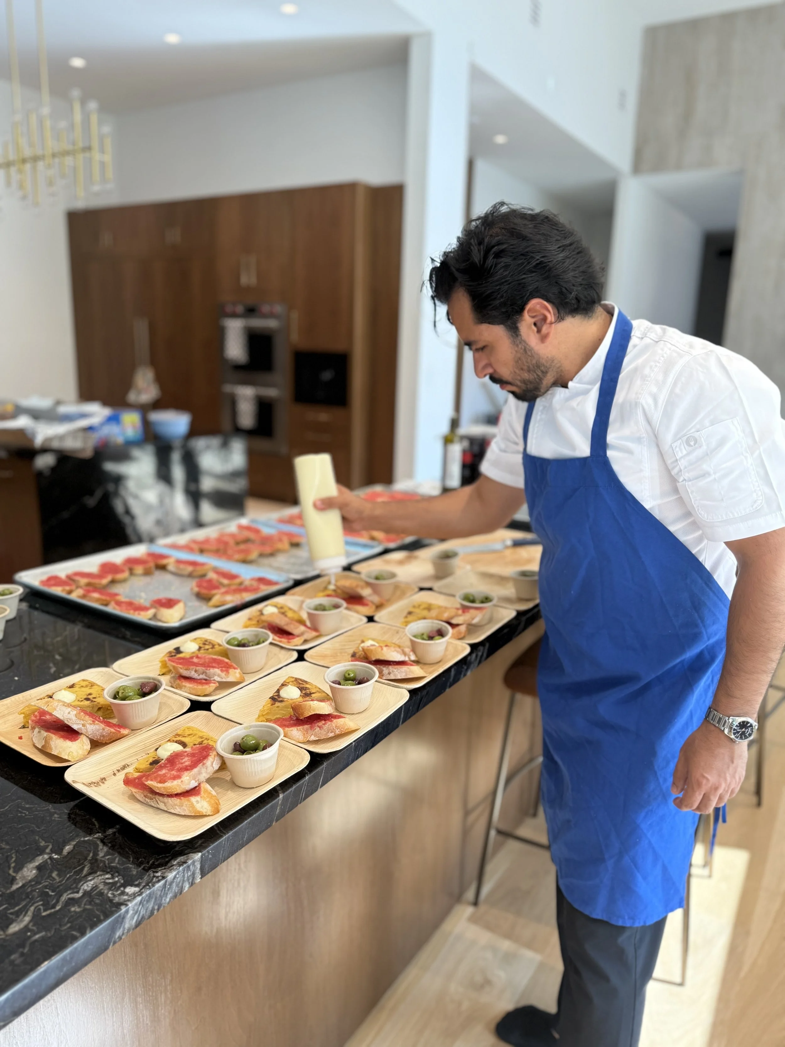 Chef Diego Cerdan wearing a white shirt and blue apron is preparing small appetizer plates with bread, sliced meat, cheese, olives, and dips on a black marble counter in a modern kitchen.