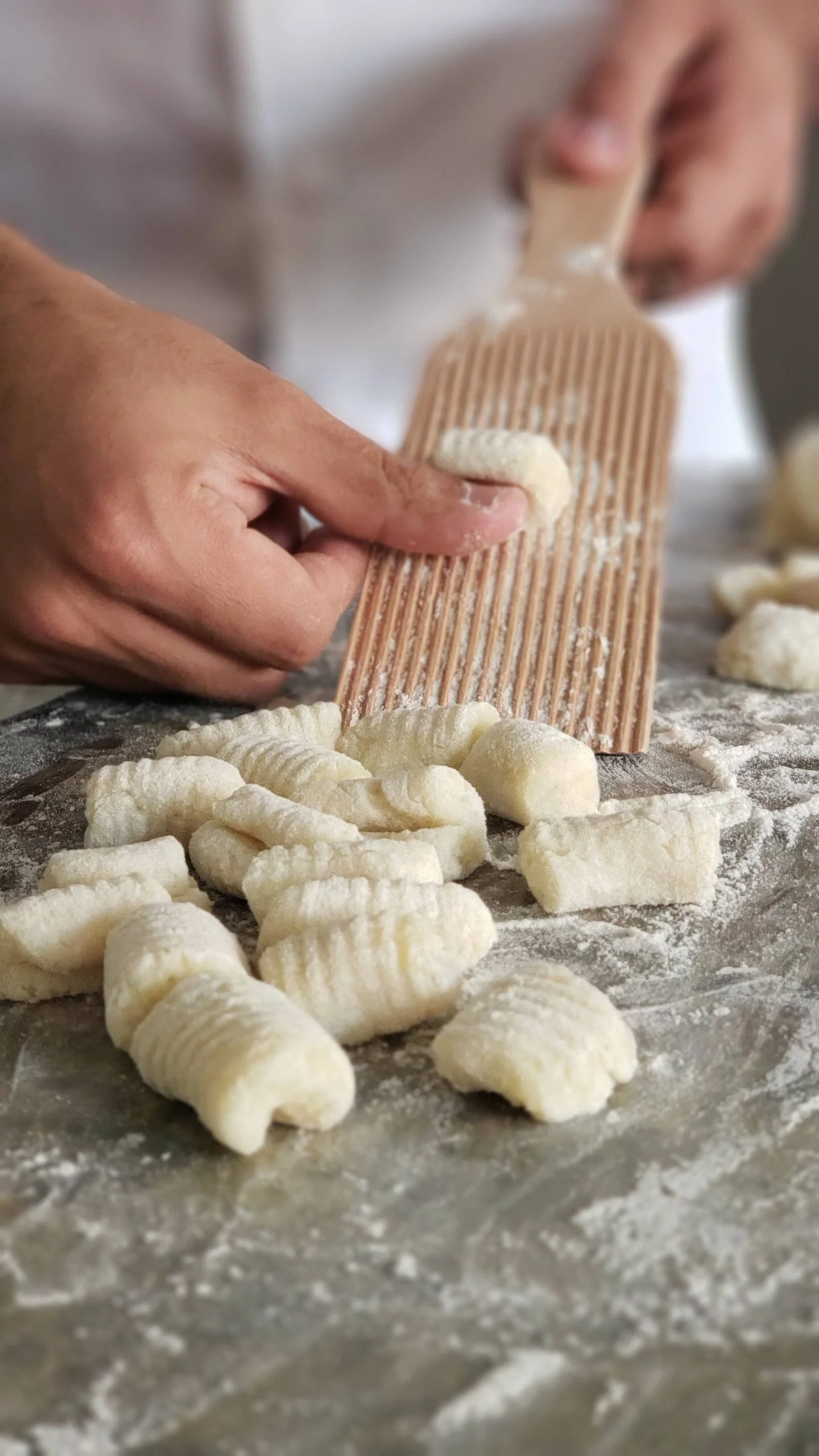 Close-up of hands preparing gnocchi dough, using a floured fork to shape the dough, with pieces of gnocchi on a dusted surface.