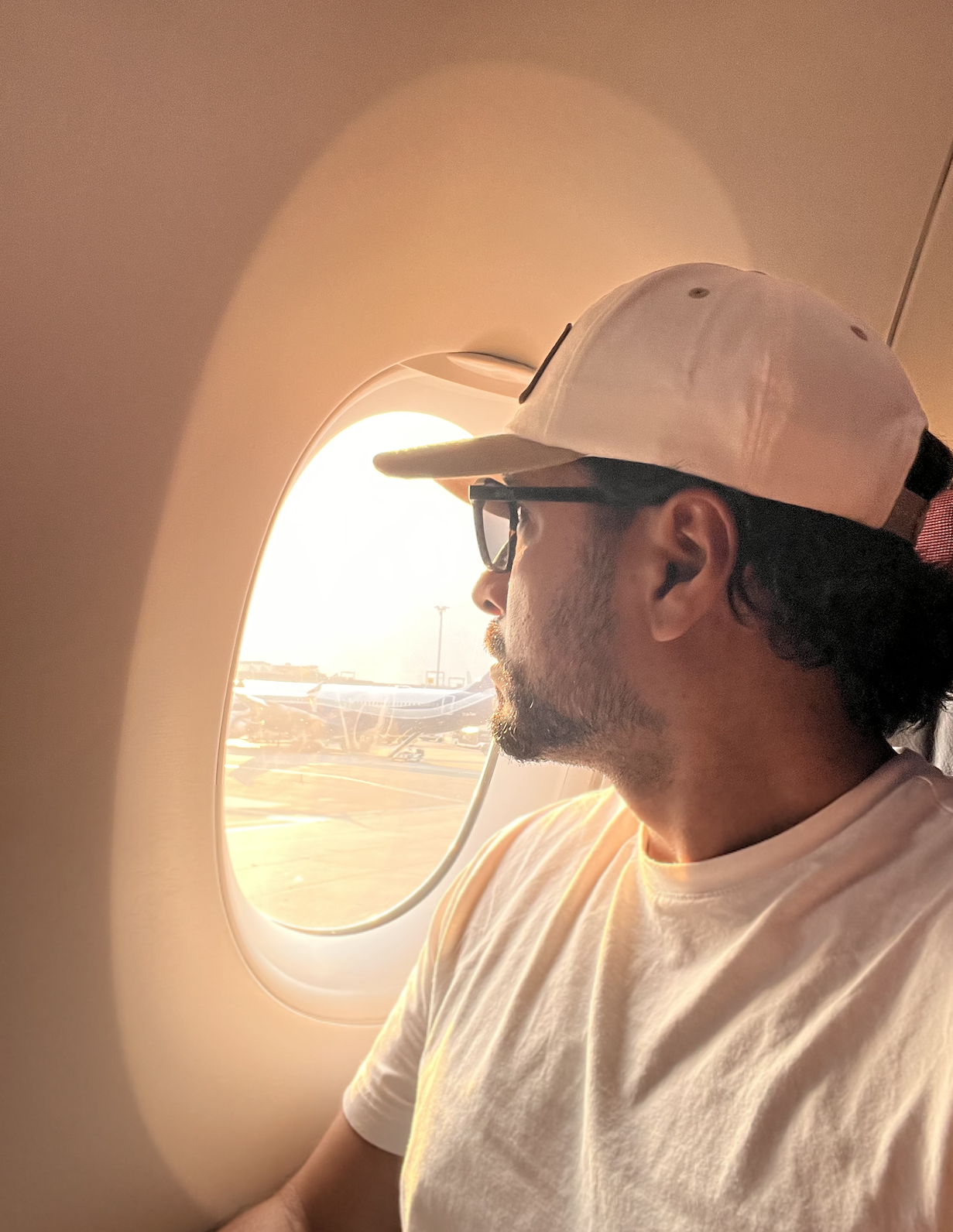 A man with glasses and a white cap, sitting in an airplane seat, looking out the window at sunset. The airport runway and planes are visible outside.