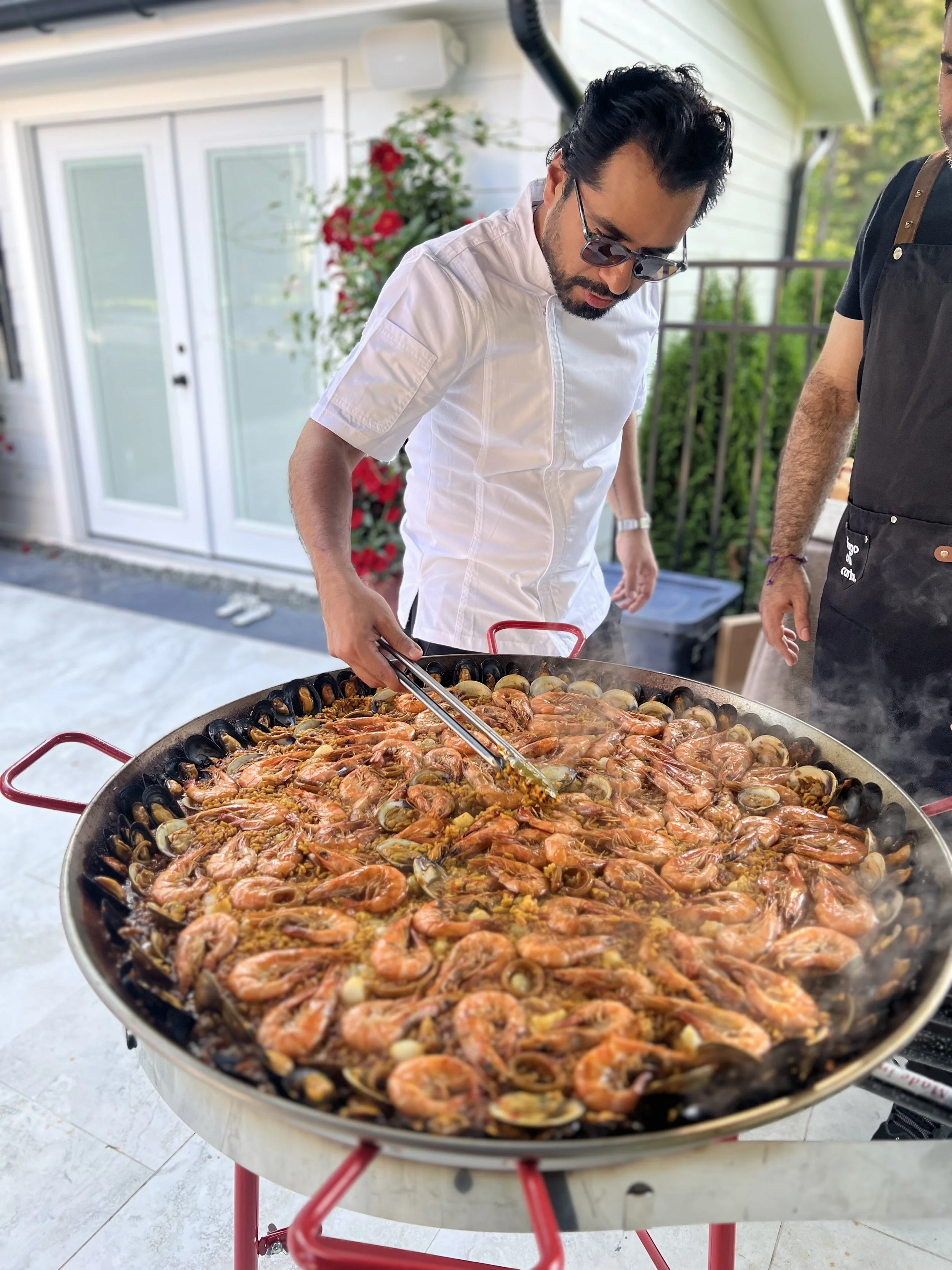Man in sunglasses and white chef coat cooking a large pan of seafood paella outdoors on a patio.
