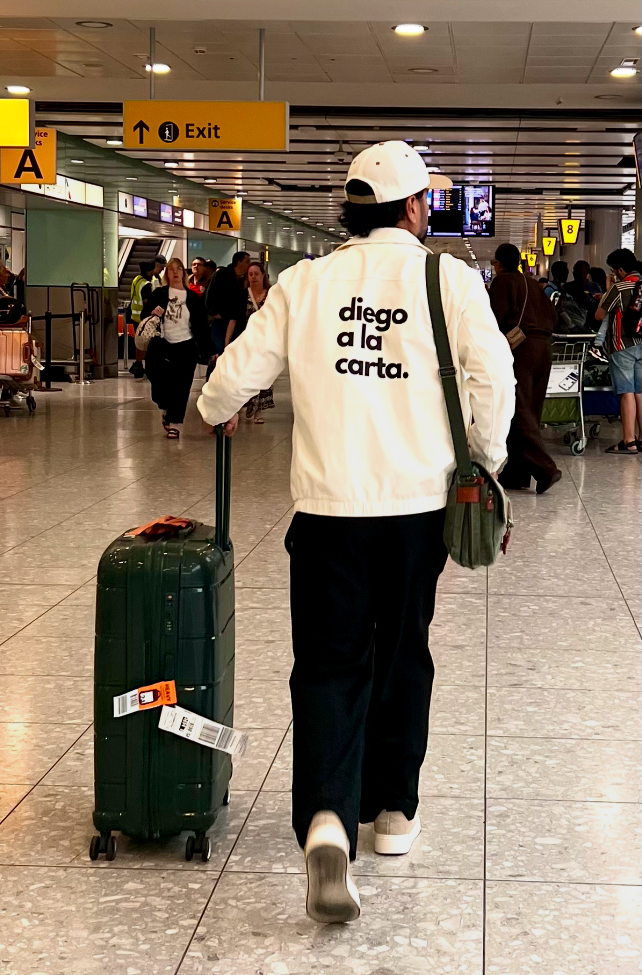 A man wearing a white jacket with the words 'diego a la carta' printed on the back, pulling a black rolling suitcase in an airport terminal.