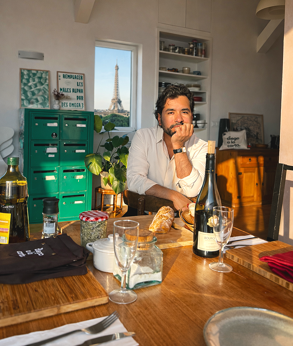 A man sitting at a dining table with a view of the Eiffel Tower in Paris, France, visible through the window. The table has food and drinks, including a bottle of sparkling wine, glasses, bread, and condiments. The background includes kitchen shelves and green mailboxes.