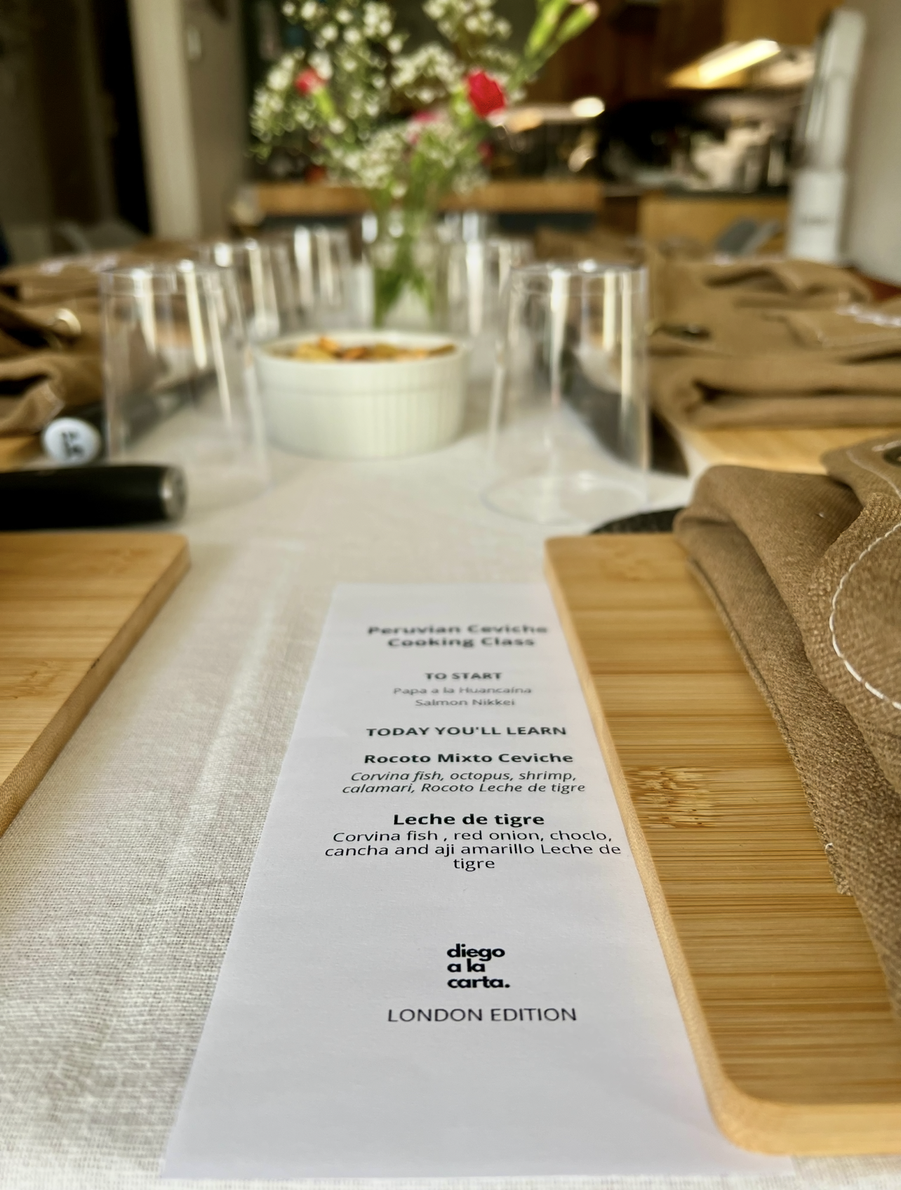 A dining table set for a cooking class with a menu, glasses, cutlery, napkins, and a centerpiece of flowers in the background.