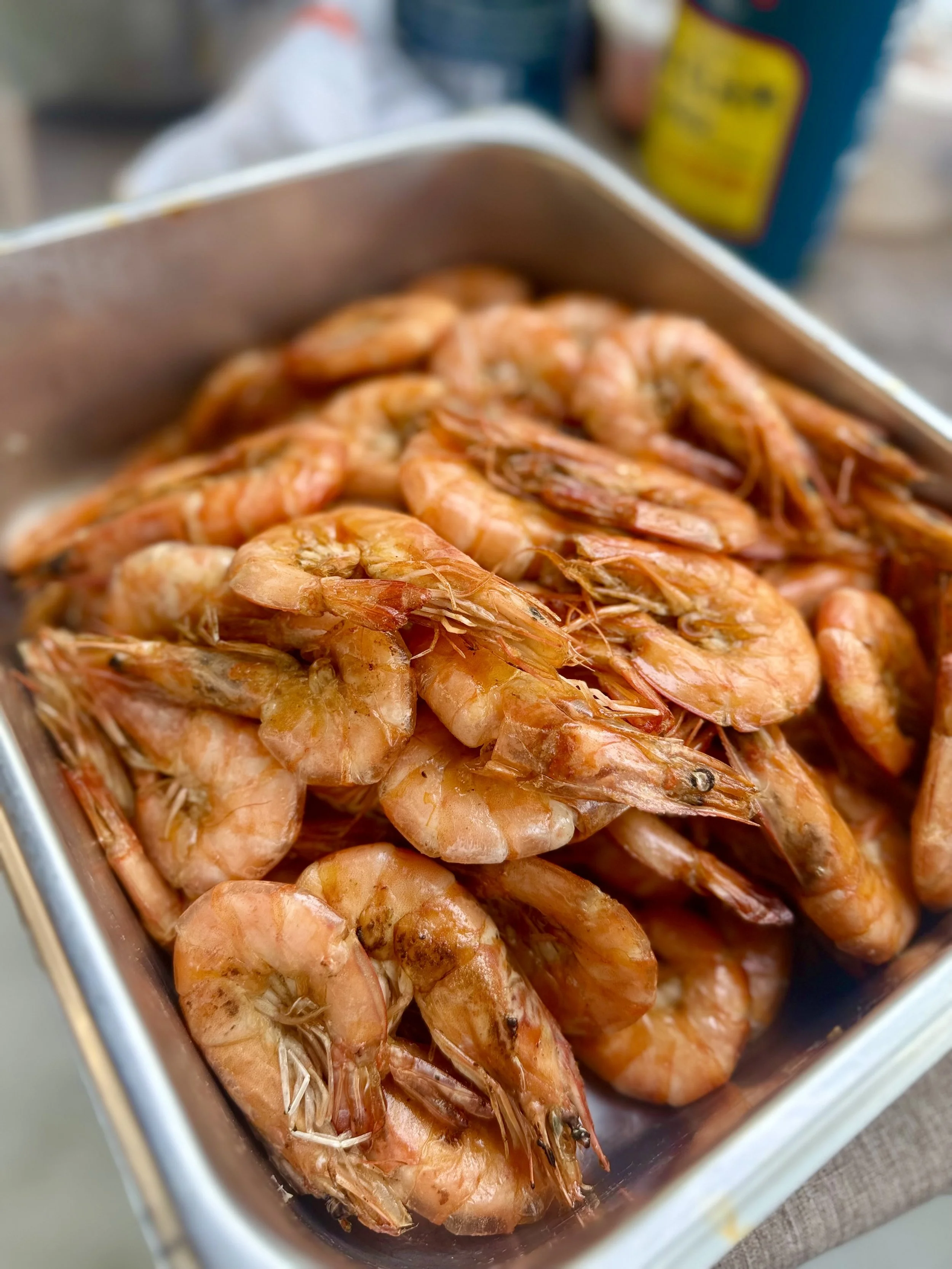 Cooked shrimp in a metal container with some blurred background including a blue and yellow container.