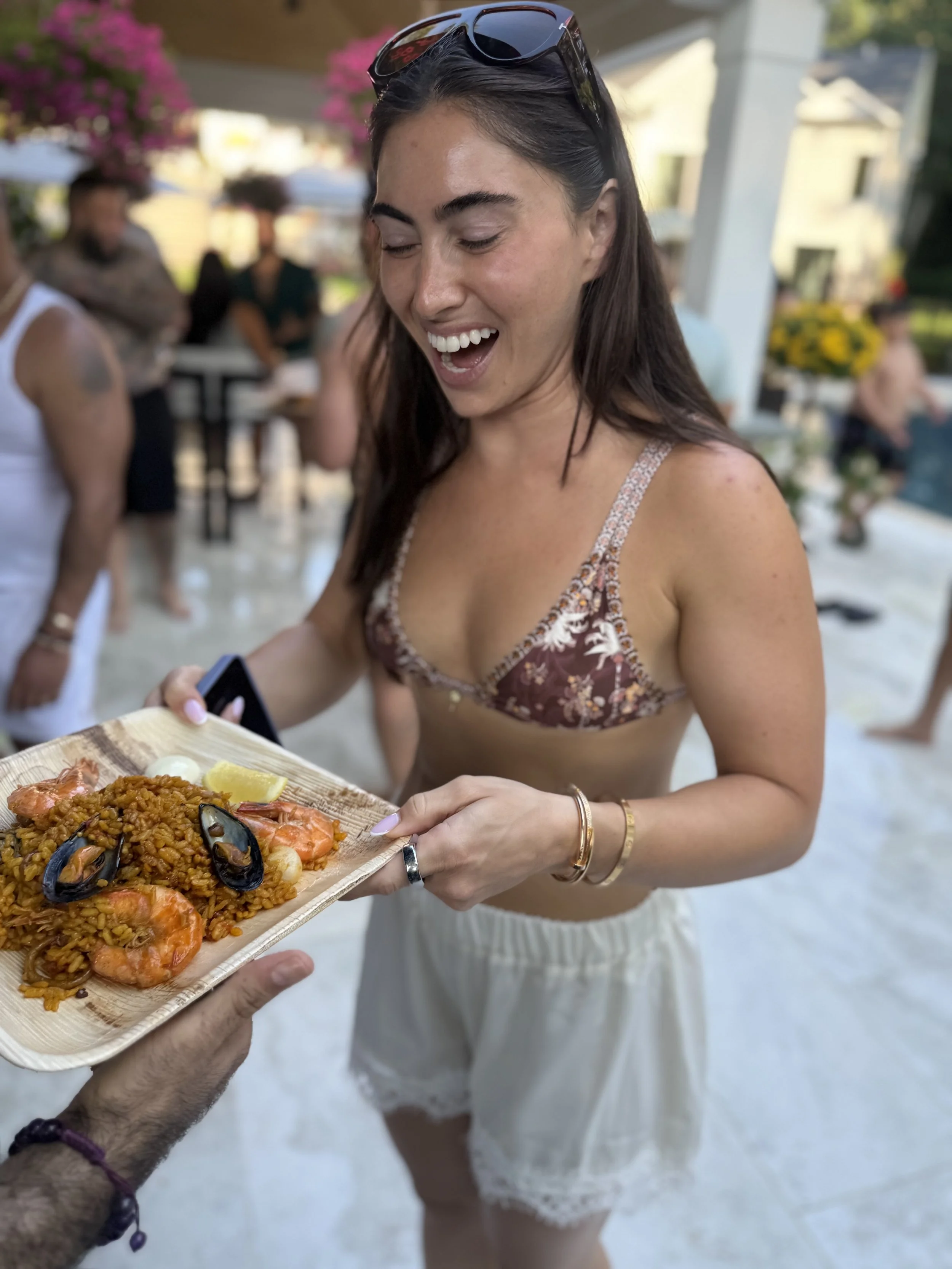 A woman in a patterned top is smiling and reaching out to receive a wooden tray of paella with shrimp, mussels, and lemon from a person in the foreground. The background shows an outdoor gathering with people, flowers, and a building.