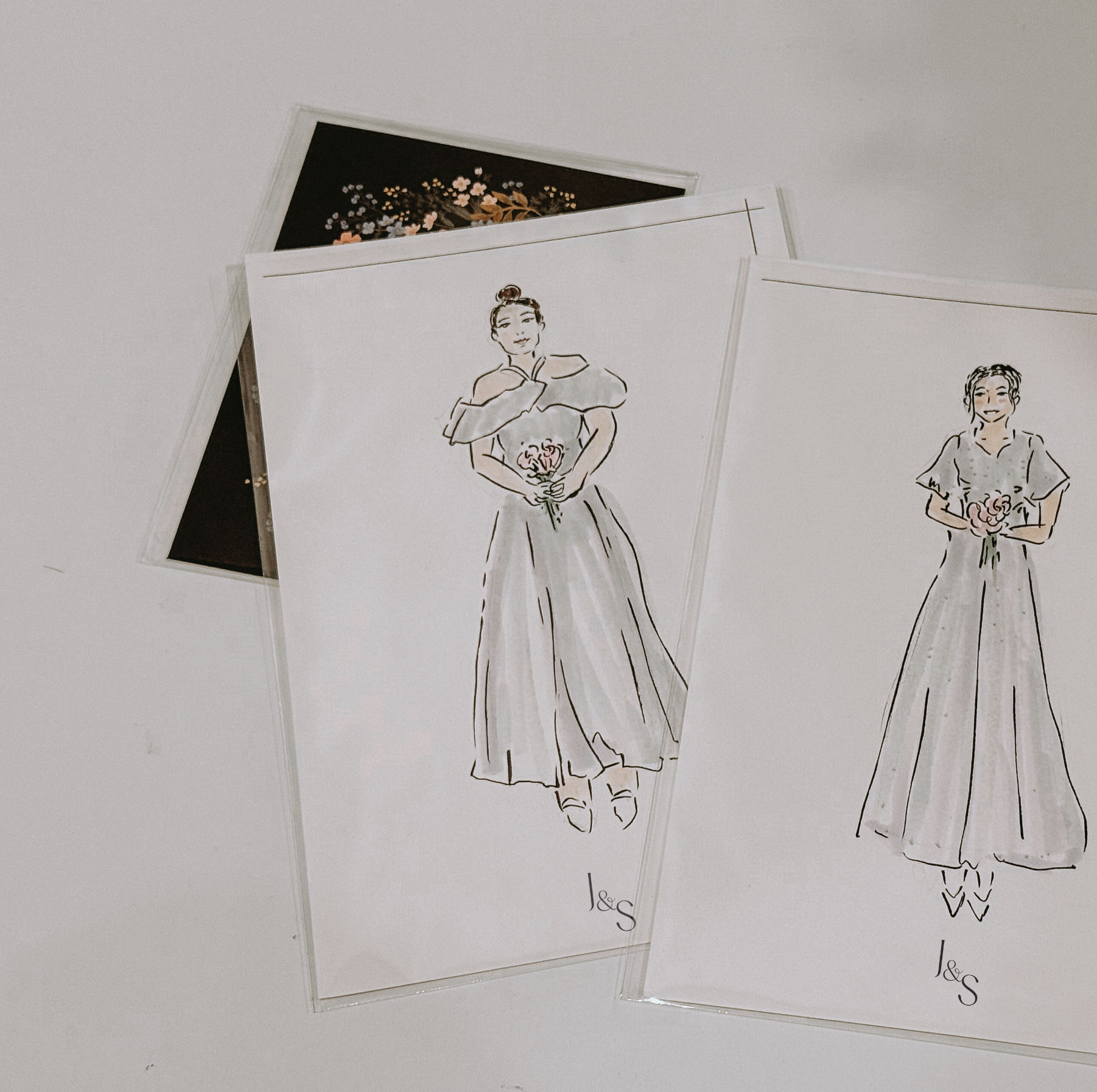 Two fashion sketches on white paper showing women in dresses, one holding flowers, with one sketch colored and the other in black and white.