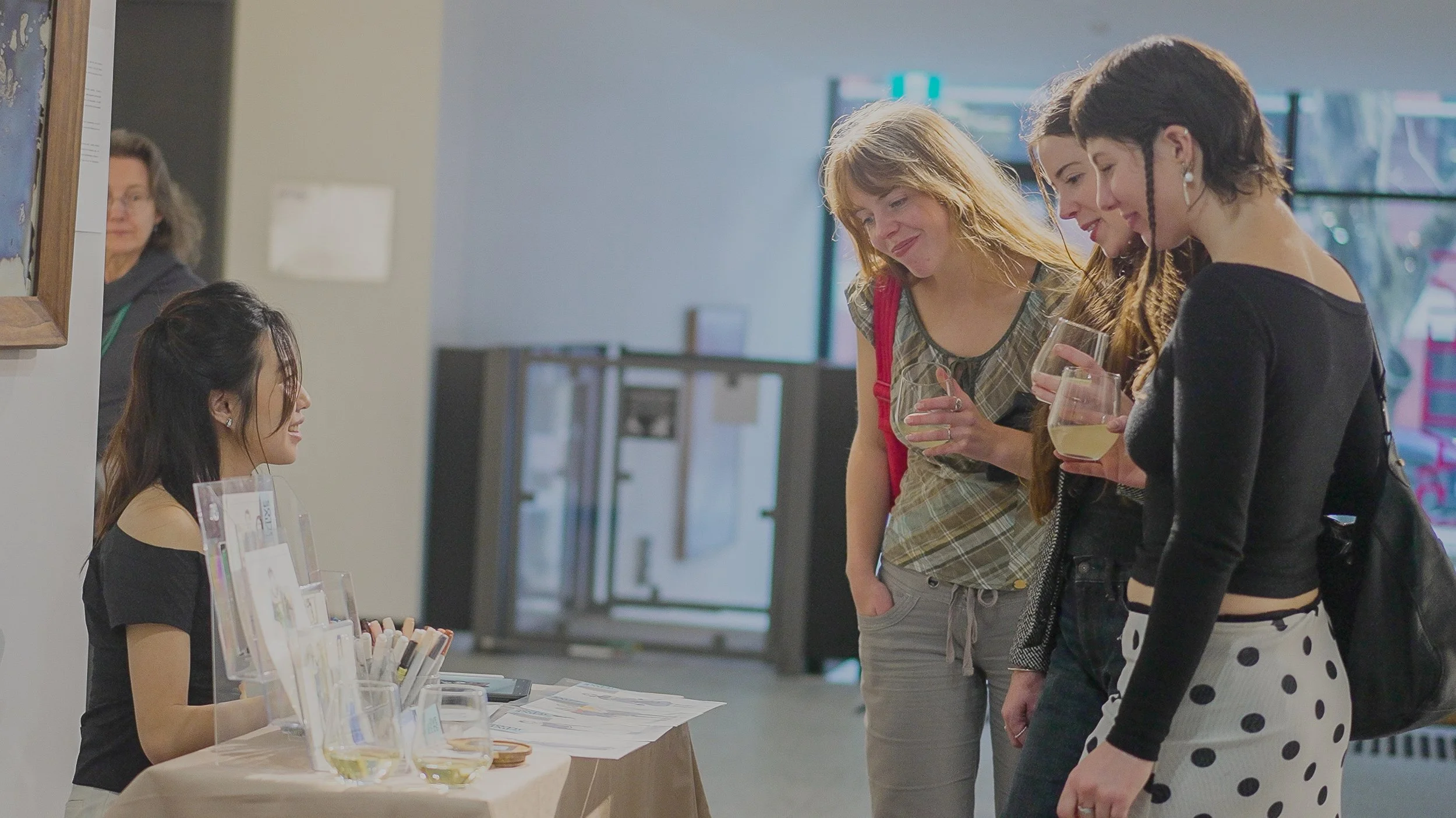 Four women at an indoor event standing near a table with brochure engaging in conversation and looking over Ives's work