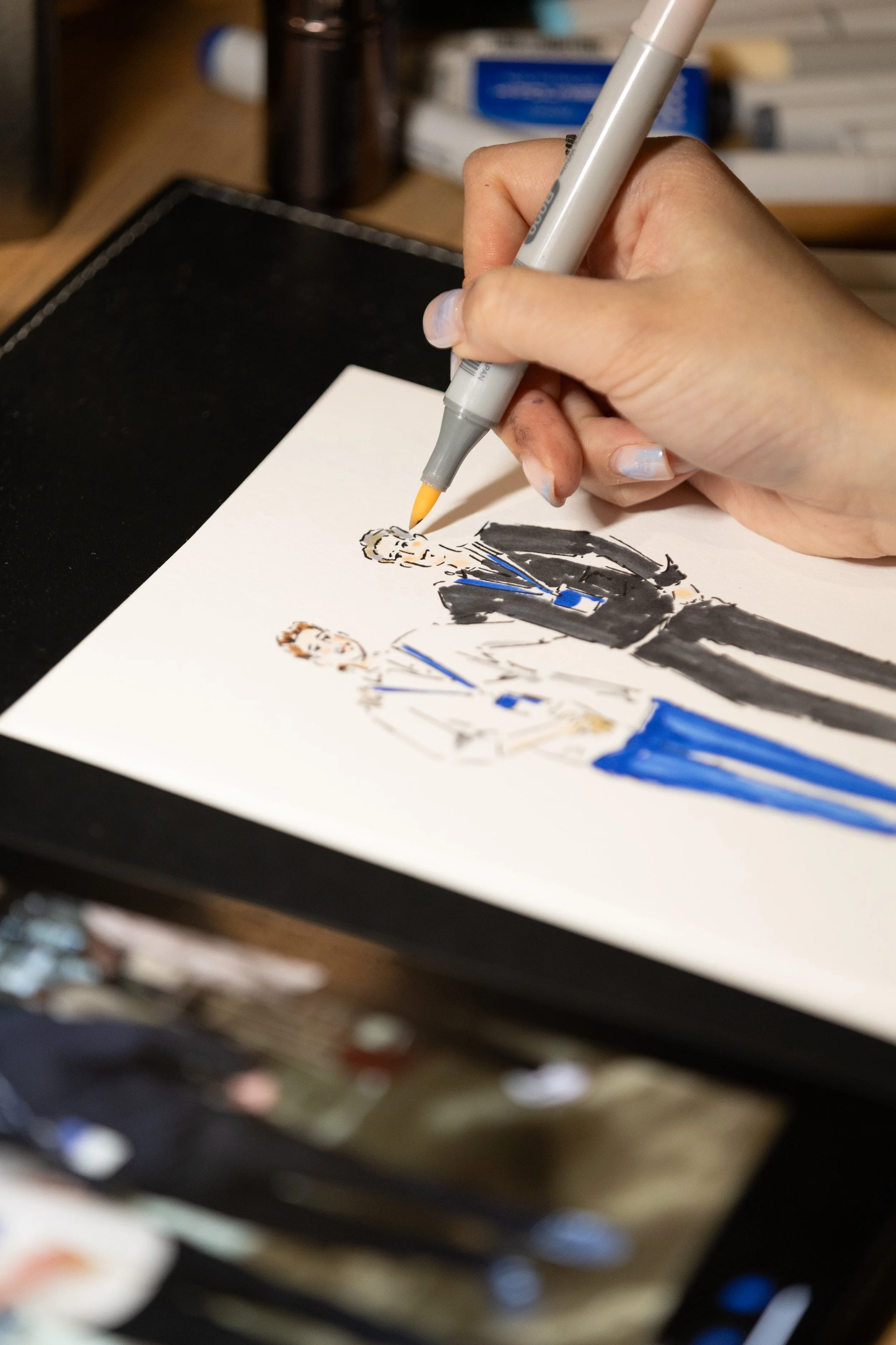 A person drawing or coloring on a white paper with a Copic marker, producing an illustration of two people in a formal setting. The background includes art supplies and a cluttered desk.