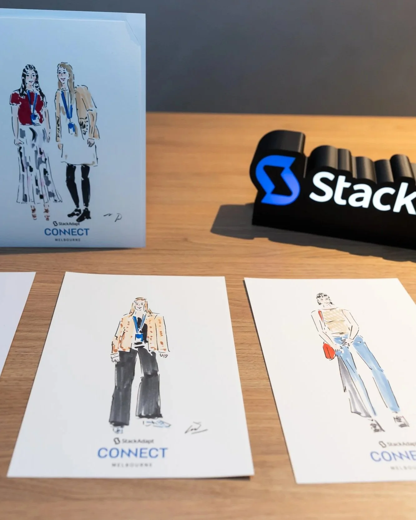 Illustrations of diverse women wearing business casual clothing with conference lanyards, displayed on a table alongside the Stack Adapt logo