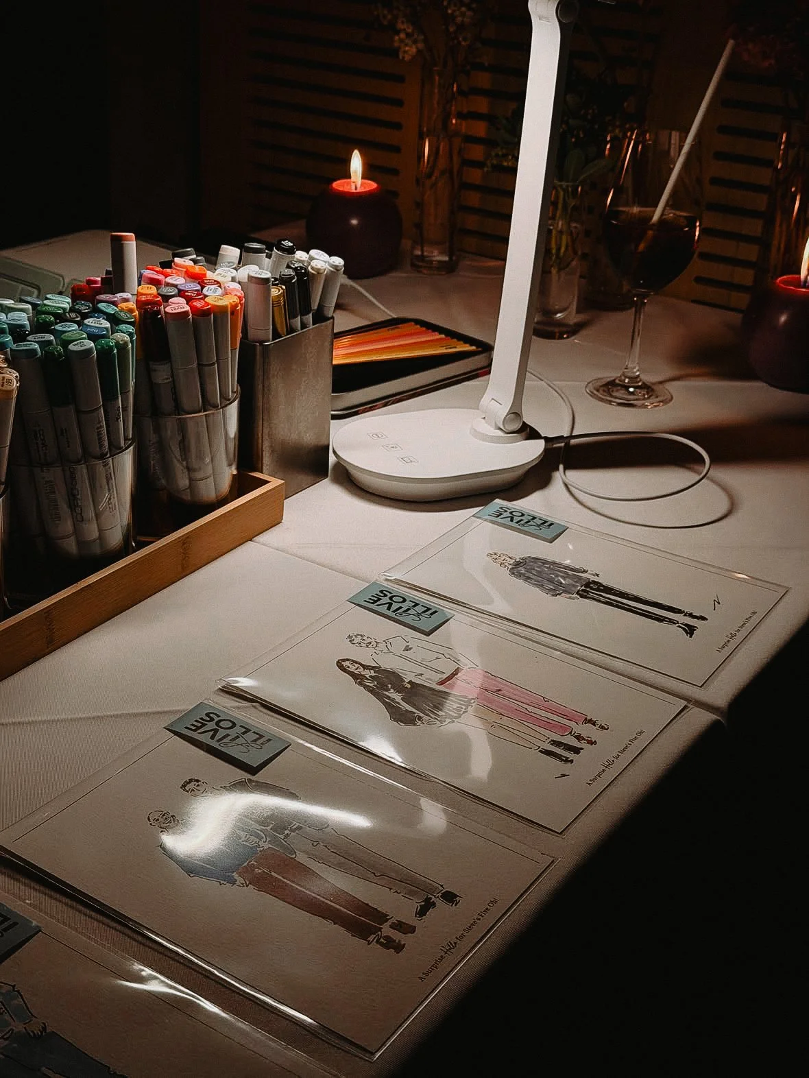 Desk setup with color markers, a lamp, a glass of drink, and fashion illustrations on paper.