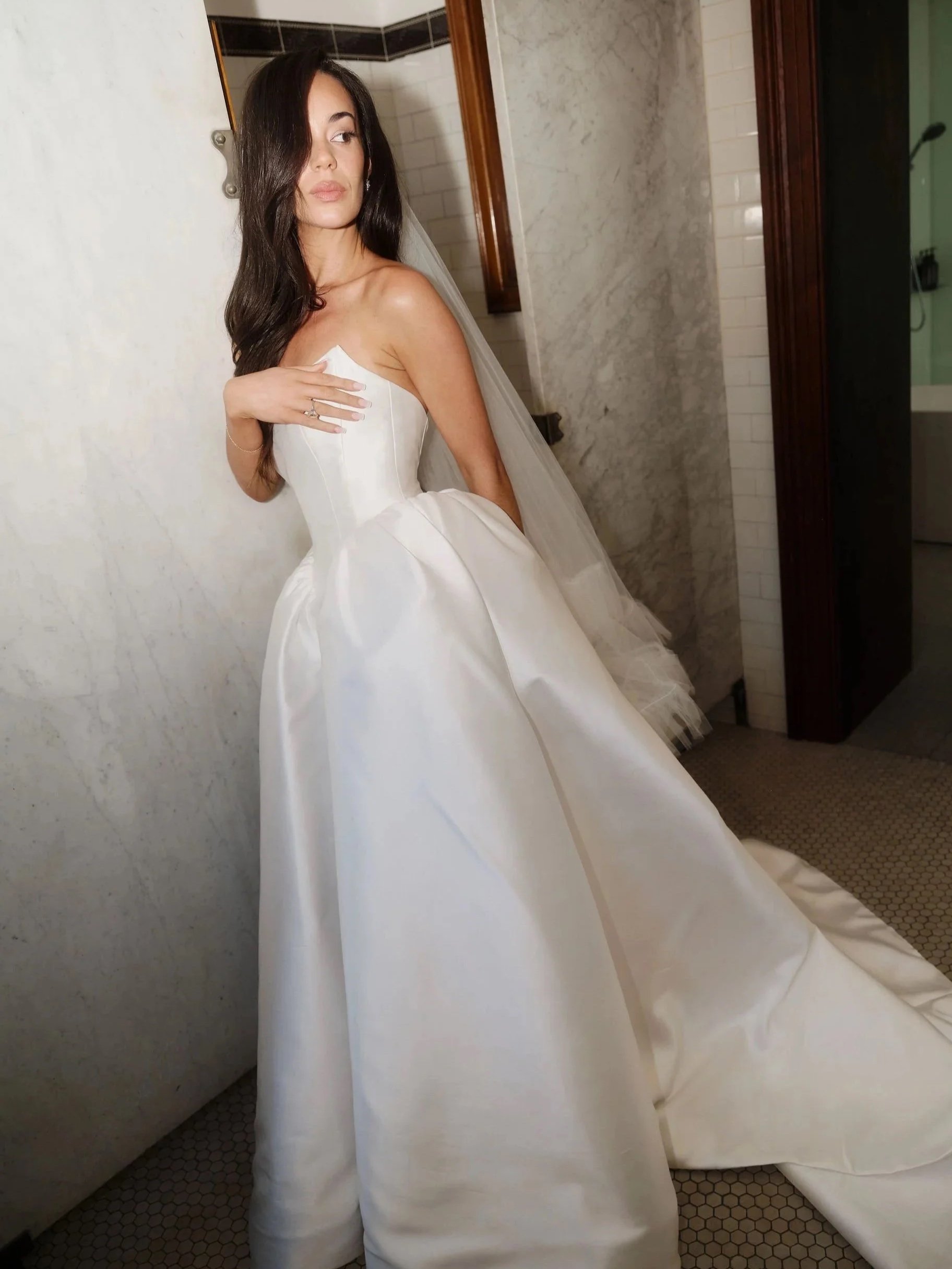 A woman in a white wedding dress with long dark hair, standing in a room with light-colored walls and a tiled floor.