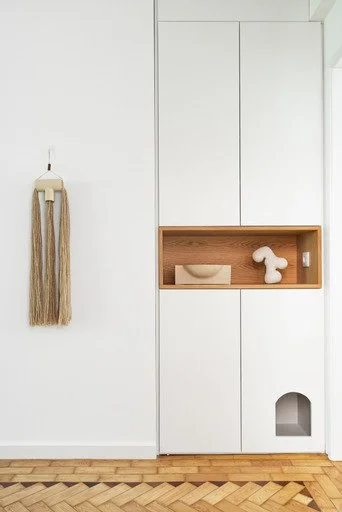 Minimalist white built-in storage with a small wooden shelf holding a decorative bowl and a ceramic figurine, next to a wall-mounted tassel decoration, on a wooden floor.